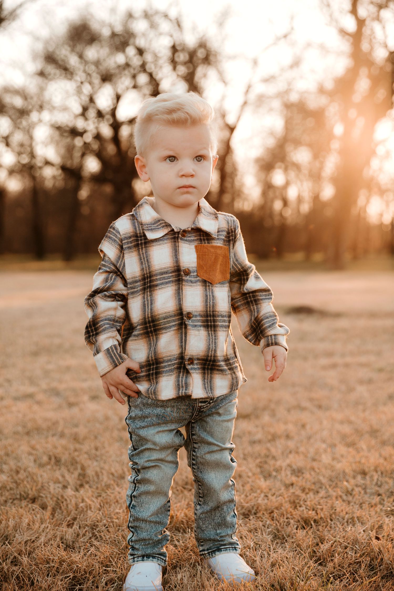 A small child wearing a plaid flannel shirt and jeans stands in a grassy field at sunset.