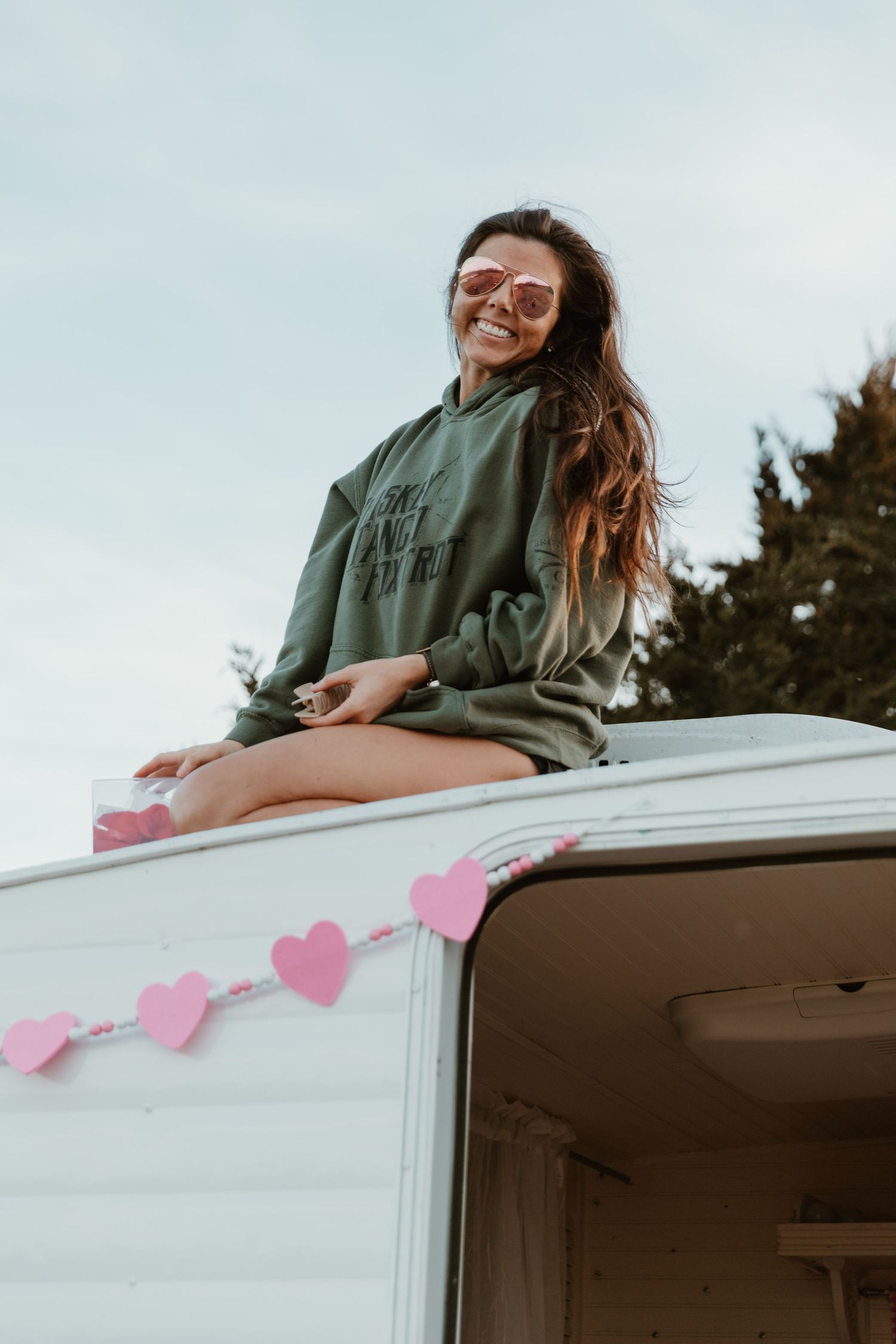 Person in green sweater sits atop white camper decorated with pink heart garland.