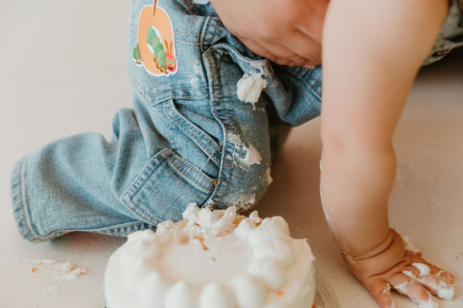 Small child in denim overalls playing with whipped cream or frosting on the floor.