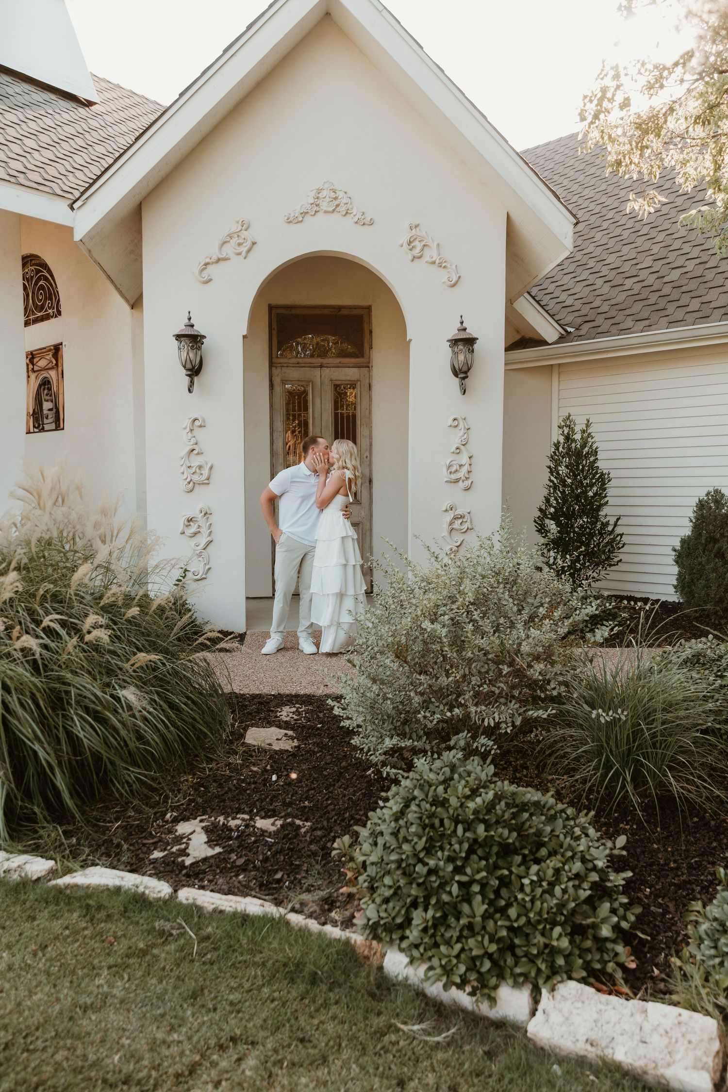 White decorative entryway with ornate moldings features lantern sconces and manicured landscaping at a residential home.