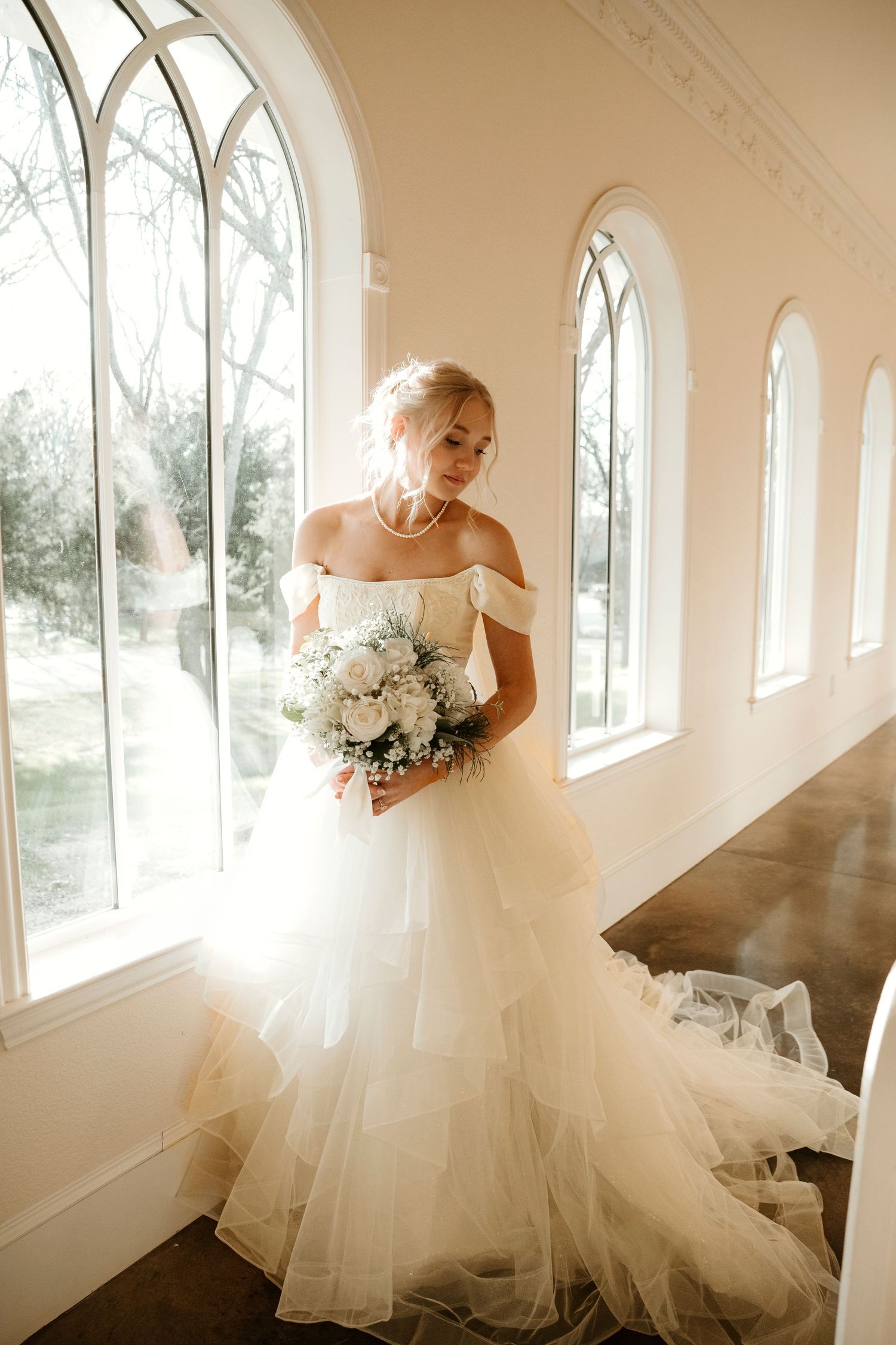 A bride in a flowing tulle gown holds a white bouquet near arched church windows.