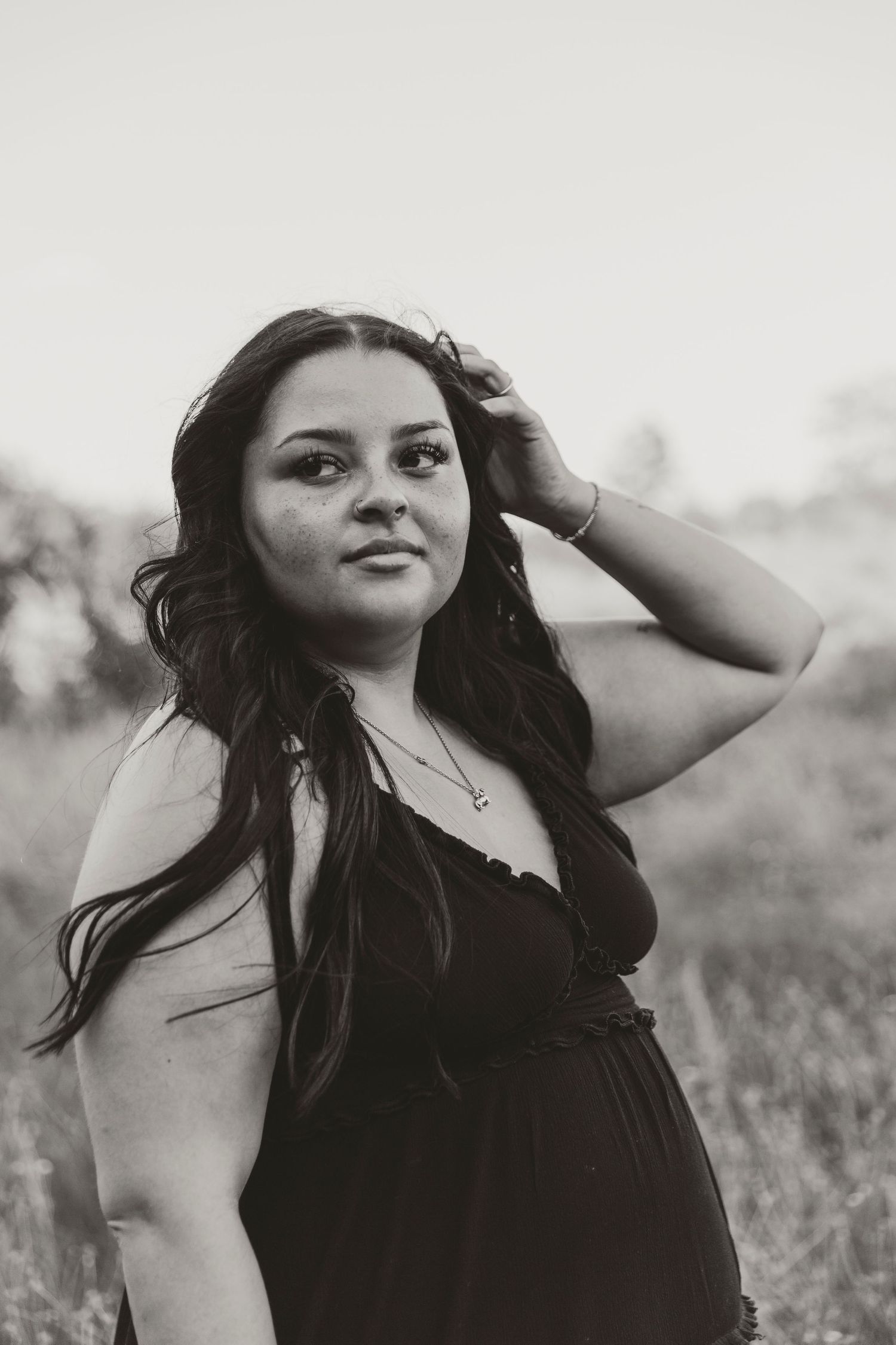 Black and white portrait photo of a woman in a dark dress with long dark hair outdoors in a natural field setting.