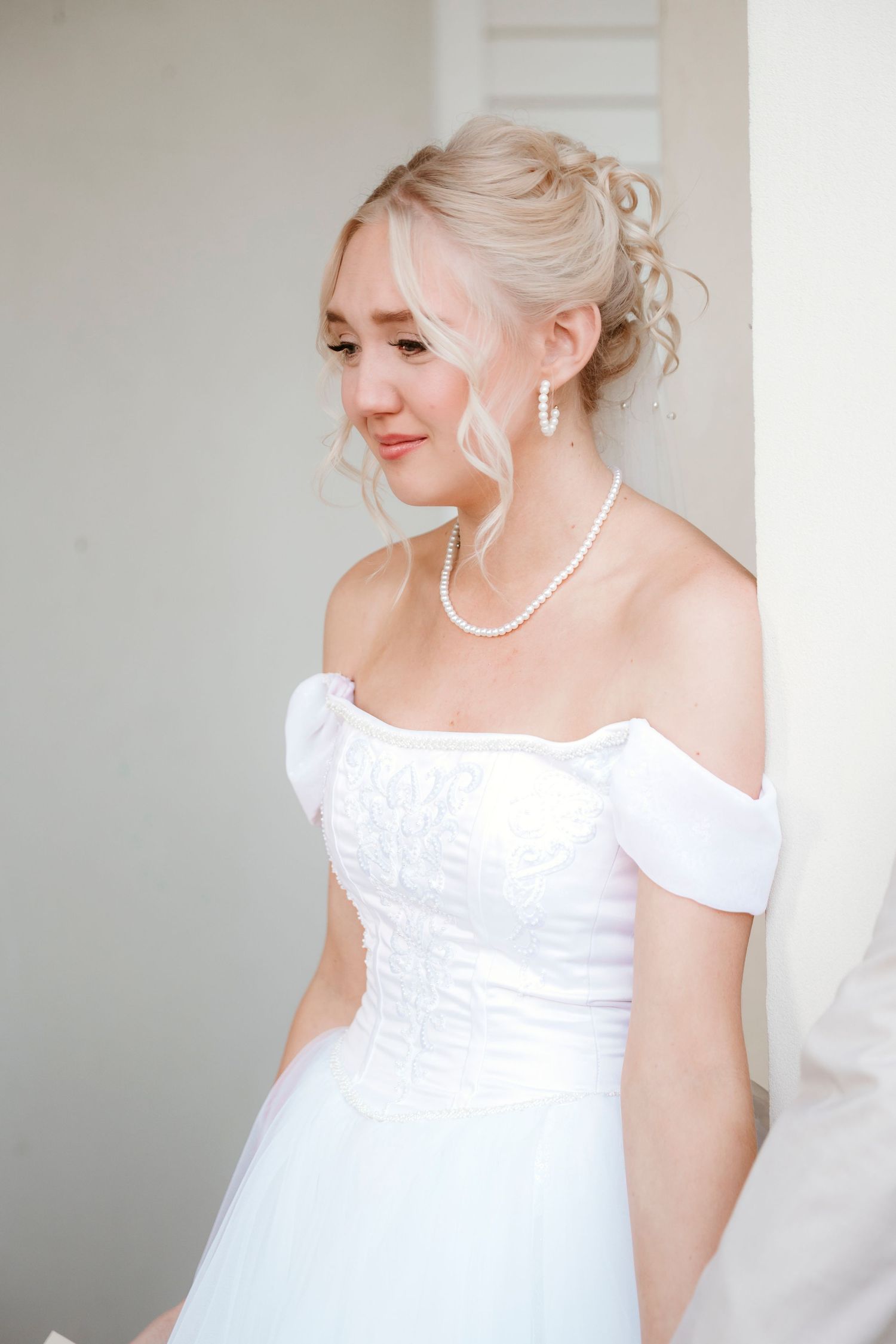 A bride in an off-shoulder white gown and pearl necklace smiles elegantly.