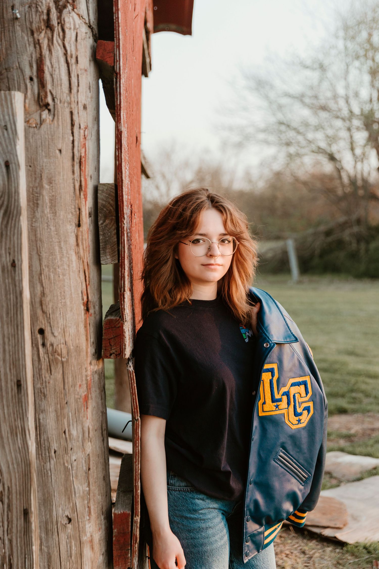 A casual outfit featuring a letterman jacket with LC letters and jeans against a rustic barn wall.
