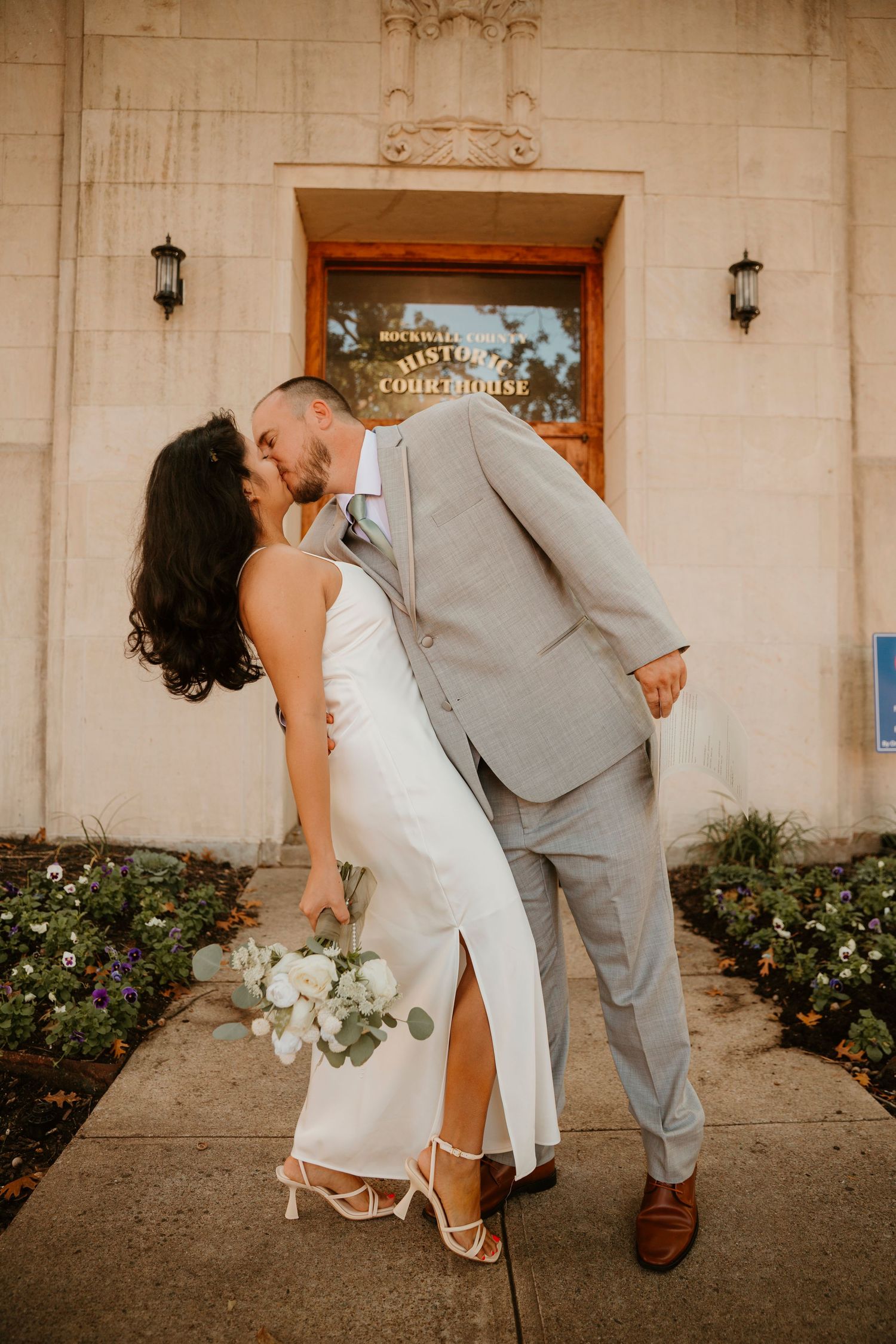 A romantic wedding couple dips and kisses in front of a stone building with flowers in the background.
