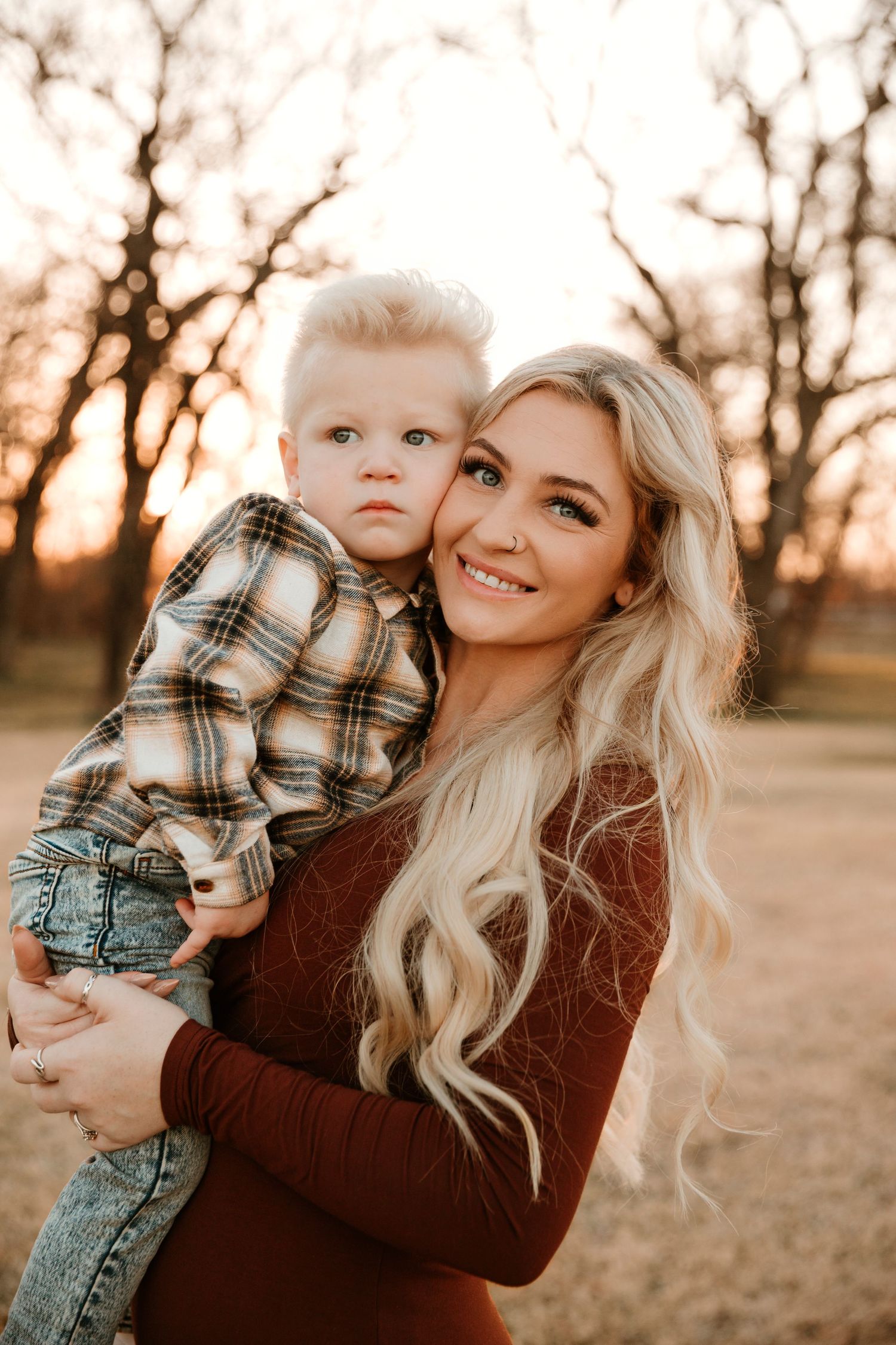 Mother and child in autumn setting with warm sunset light behind bare trees wearing plaid and burgundy.