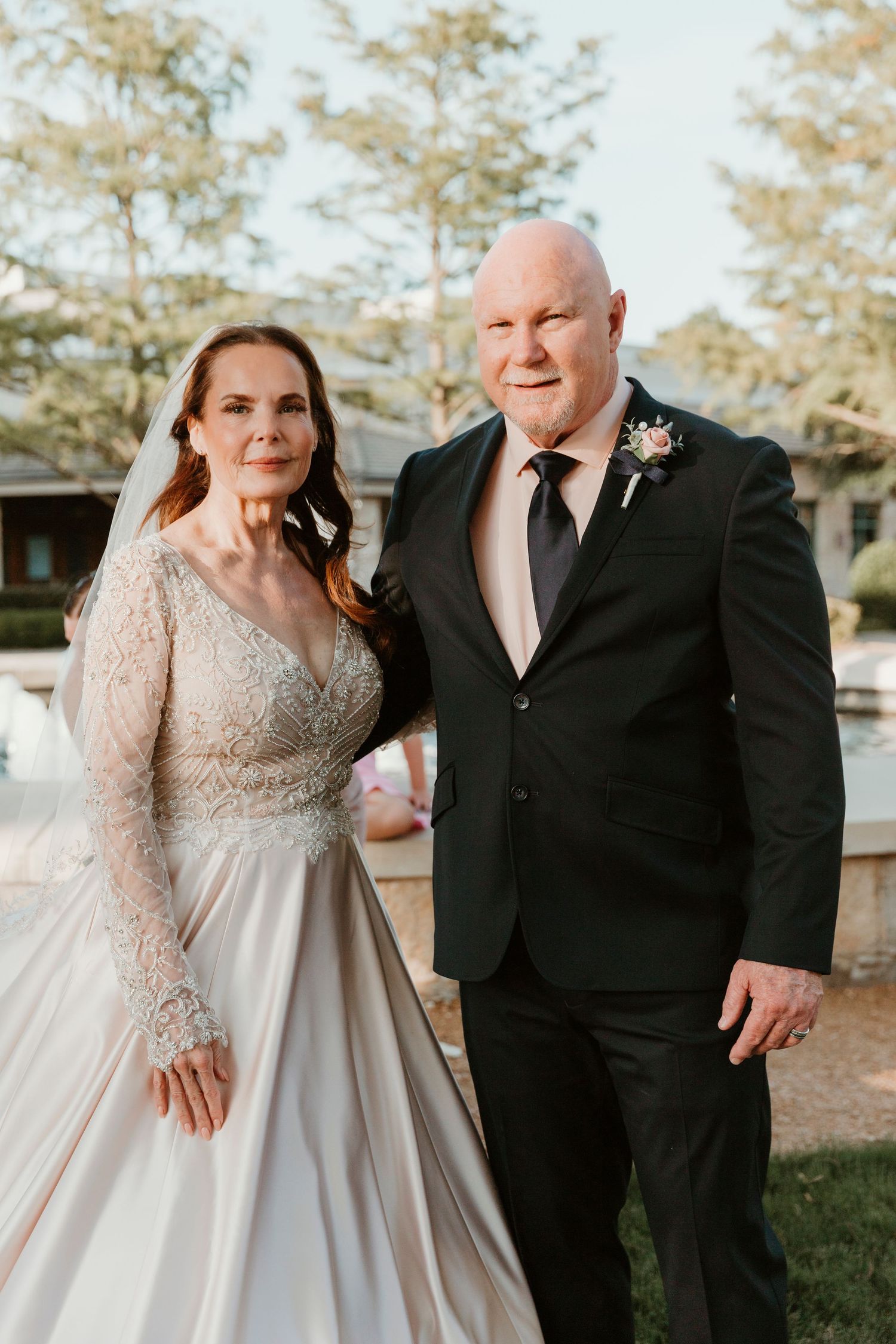 Elegant bride in lace-sleeved wedding gown poses with groom in black suit and blush tie at outdoor venue.