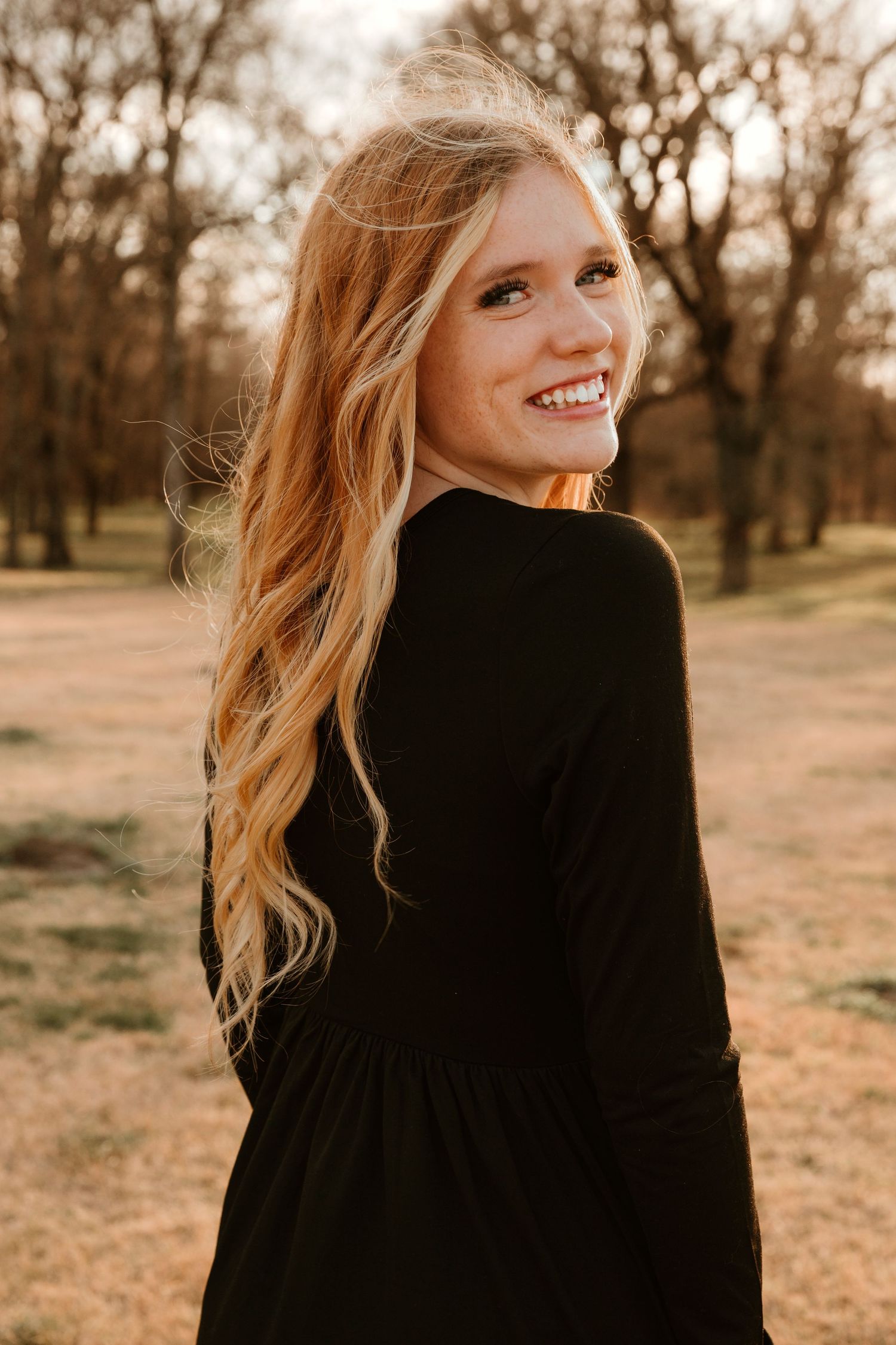 Portrait of a woman in a black sweater smiling over her shoulder at sunset in a wooded park setting.
