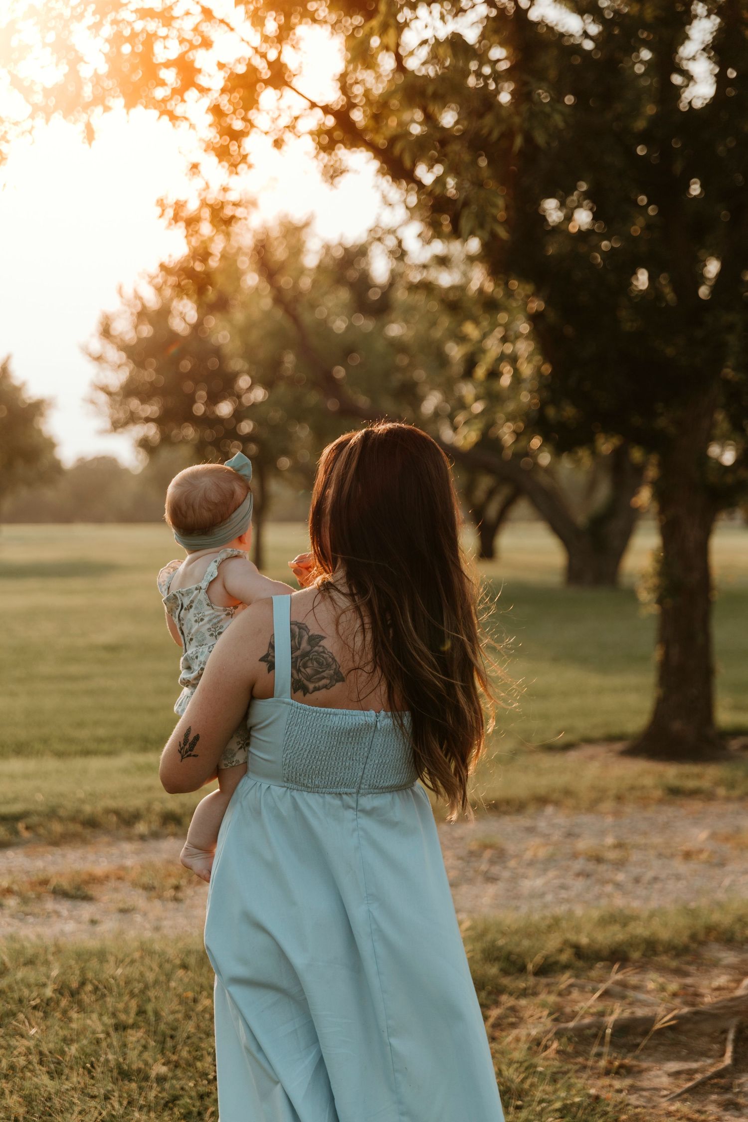 Person in a light blue dress holding a small child while walking through a sunset-lit park.