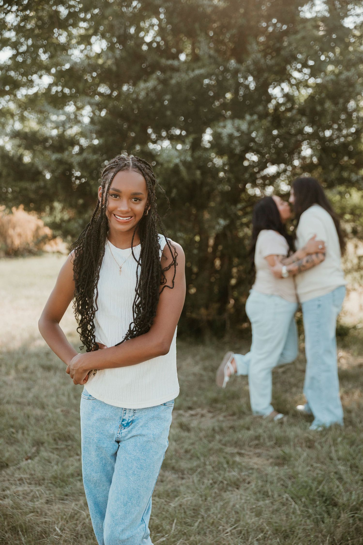 Young woman with braids smiling outdoors in white top and jeans, two friends hugging in background.