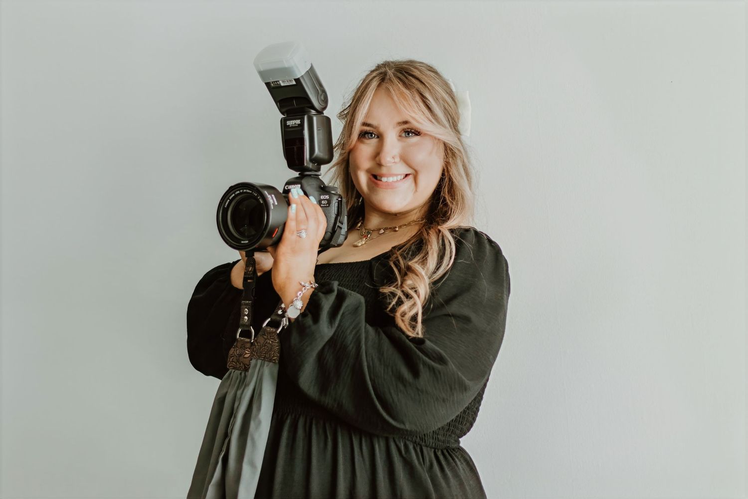 Professional photographer in black dress holding DSLR camera with flash against light background.