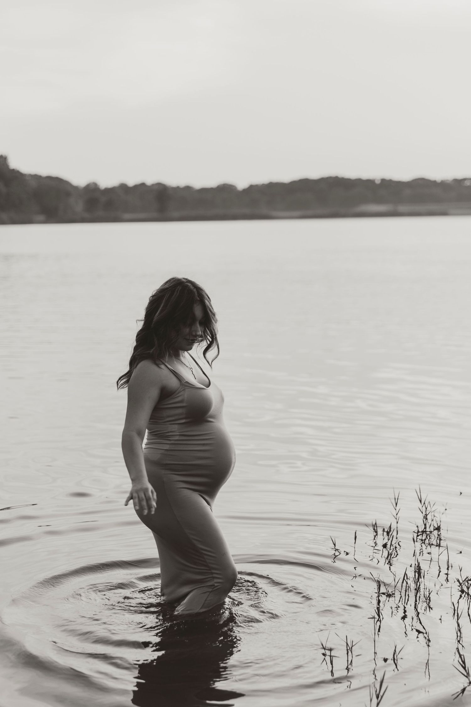 A pregnant silhouette standing in a lake at dusk creates ripples in the calm water.