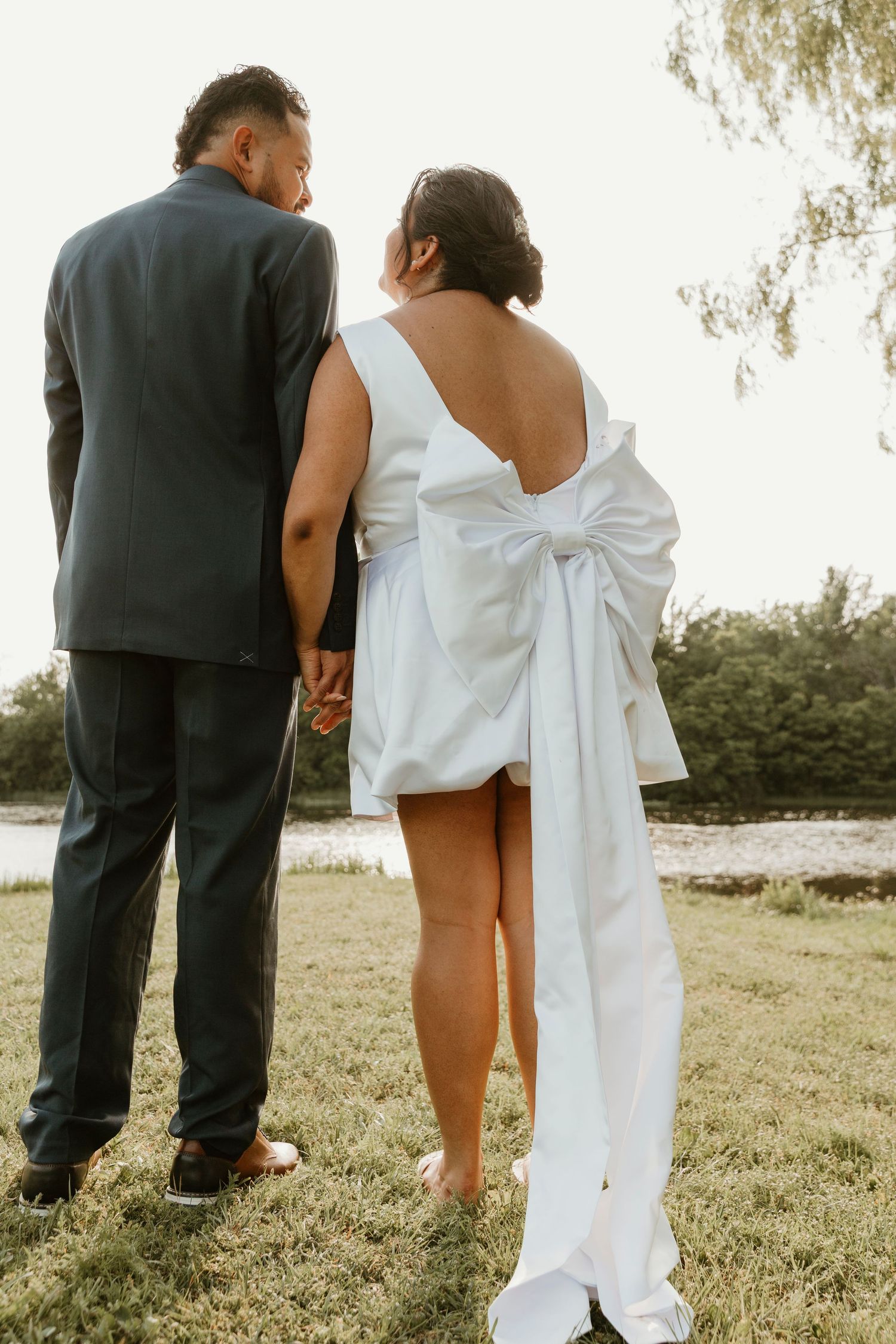 Couple standing together in grassy field with bride wearing distinctive bow dress.