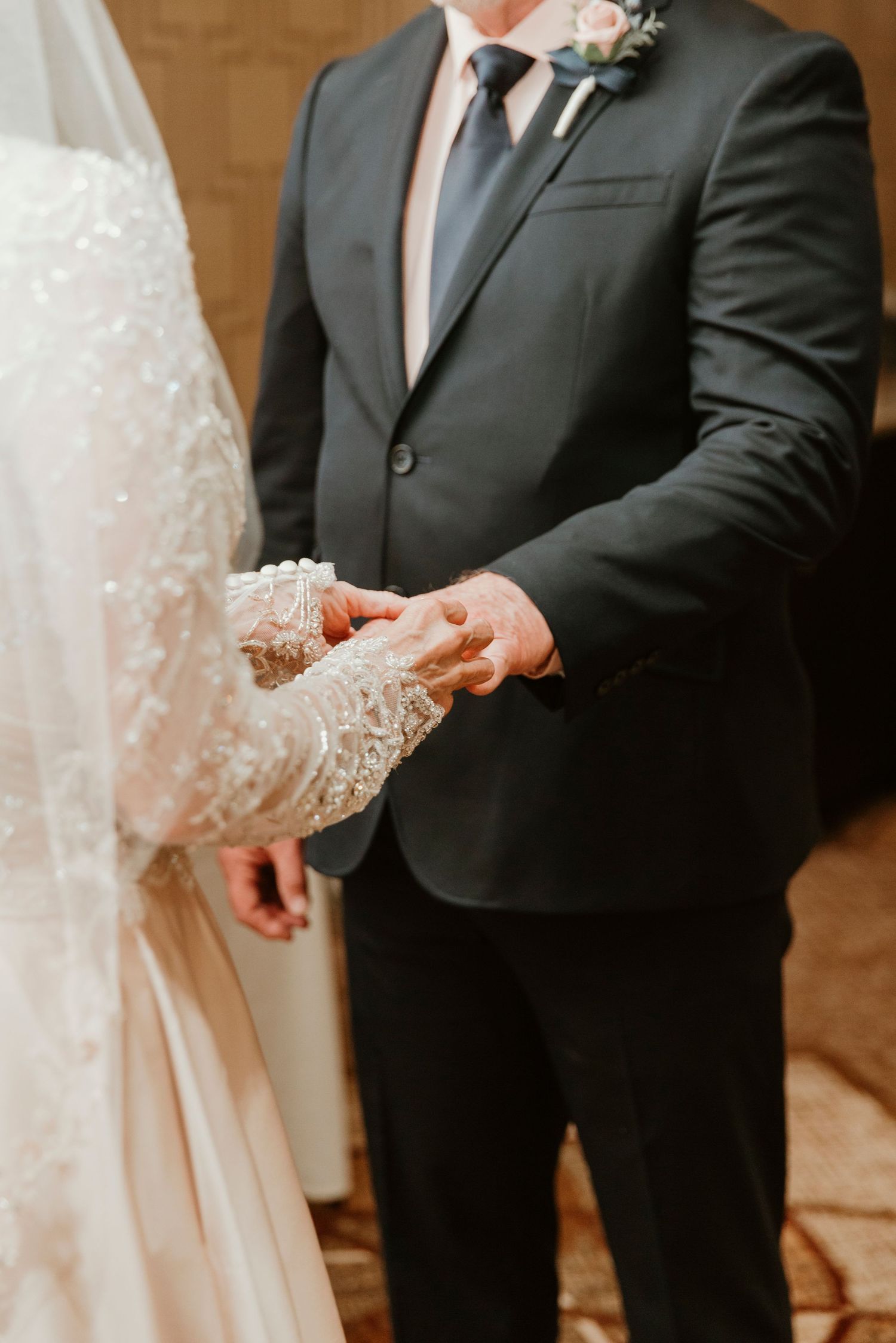 Close-up shot of bride and groom's hands during wedding ceremony, with lace dress detail and dark suit visible.