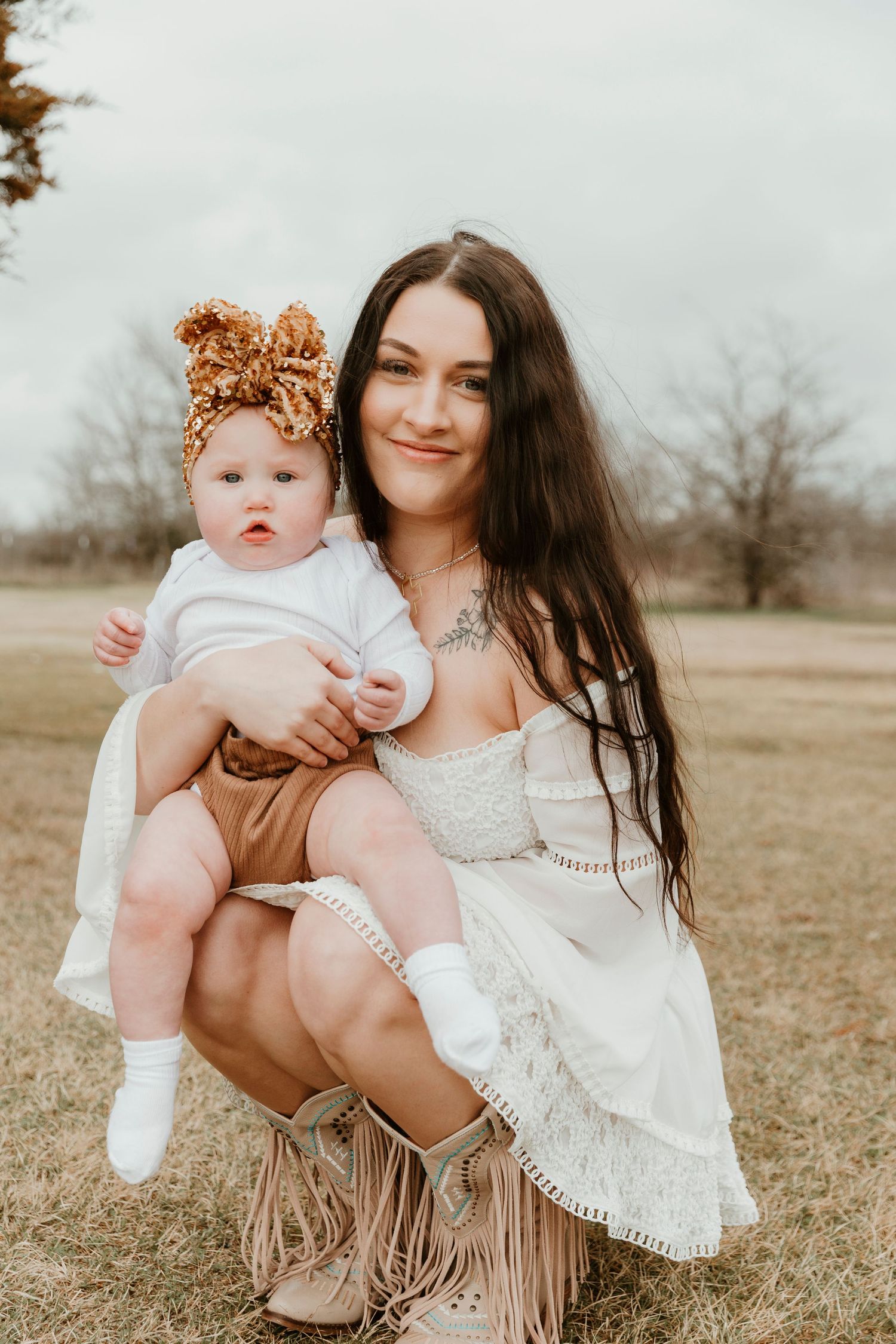 A woman in a white lace dress holding a baby wearing a brown outfit and bow headband in an outdoor rural setting.