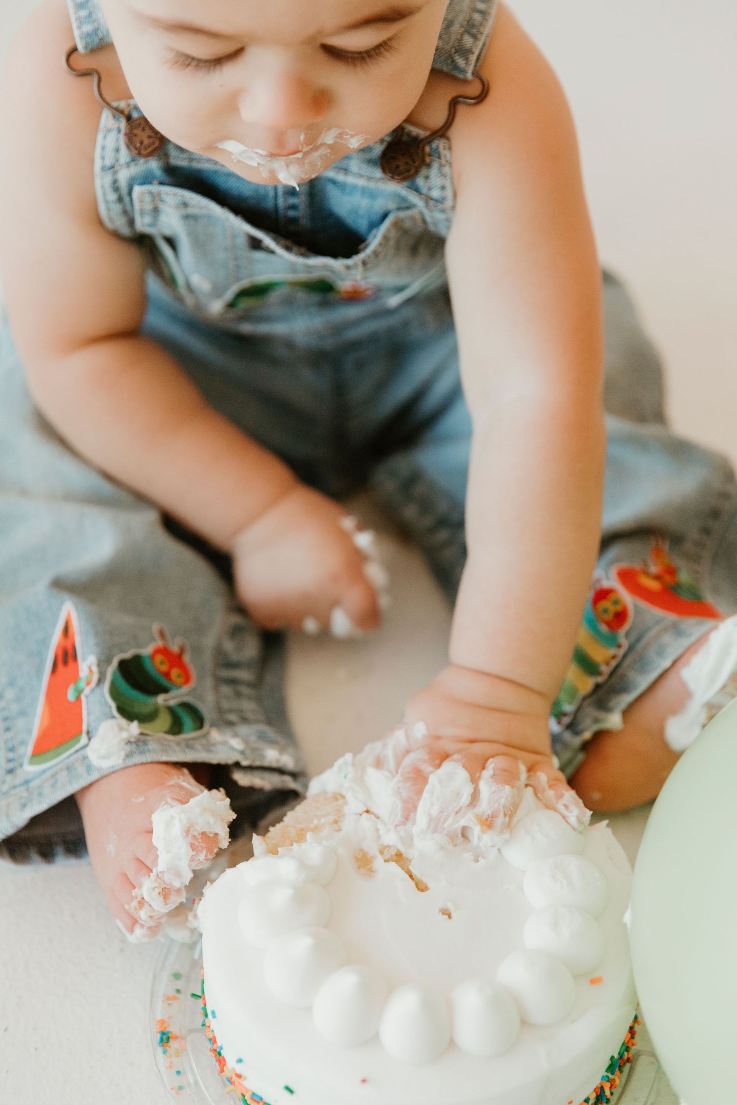 Small child in denim overalls playing with white frosting and colorful balloons on a light background.