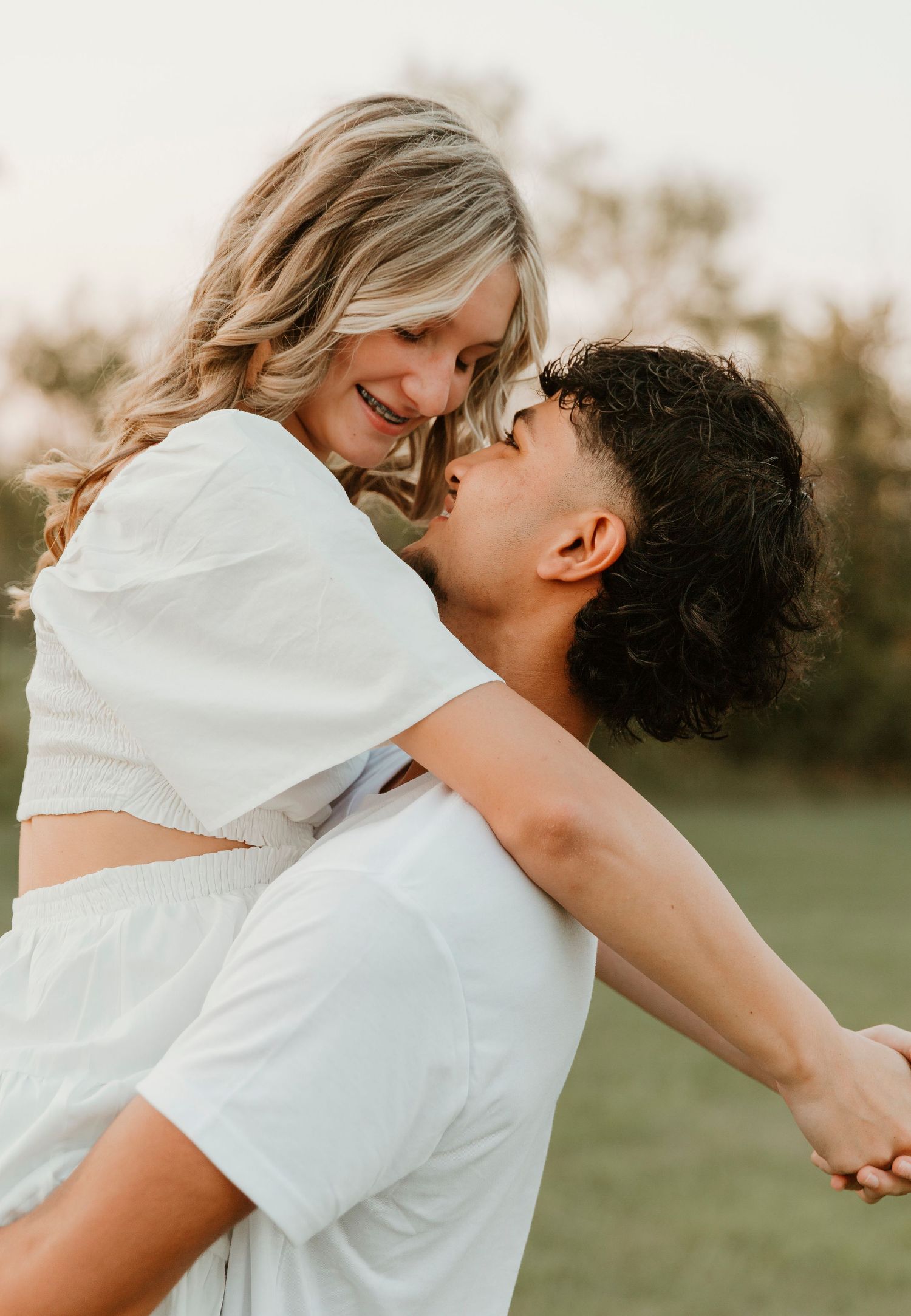 A romantic couple dressed in white embraces outdoors during a sunset photoshoot in a grassy field.