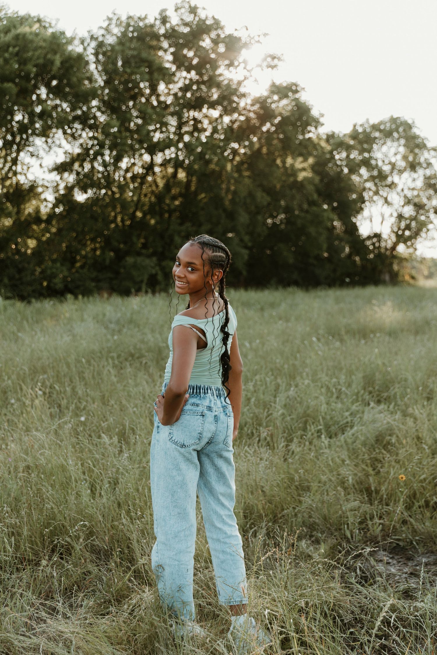 Someone wearing denim overalls and mint top stands in tall grass during golden hour.