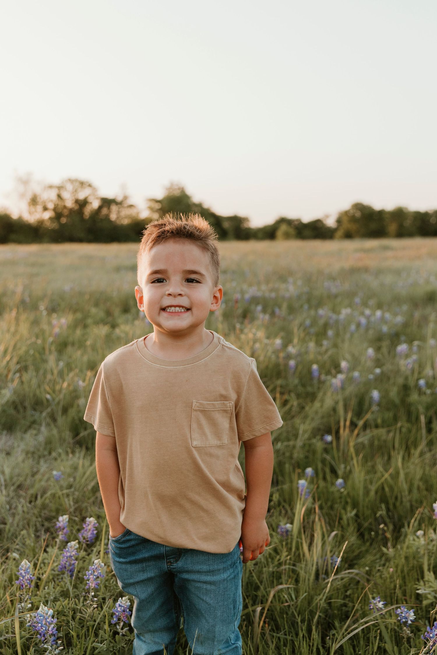 Young boy in tan shirt smiling in meadow with purple wildflowers.