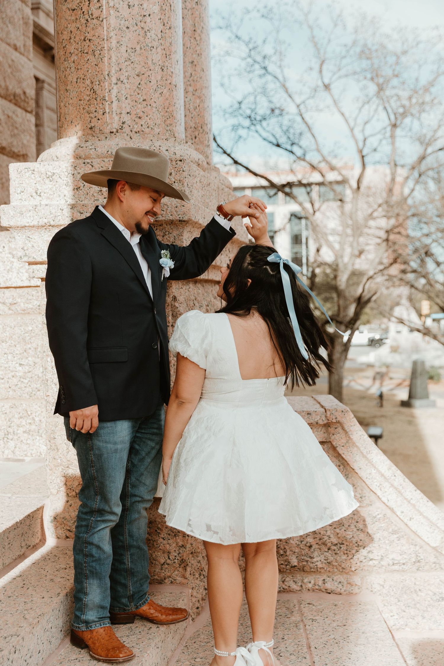 A couple in western wedding attire stands on stone steps, with the groom in cowboy hat and bride in short white dress.