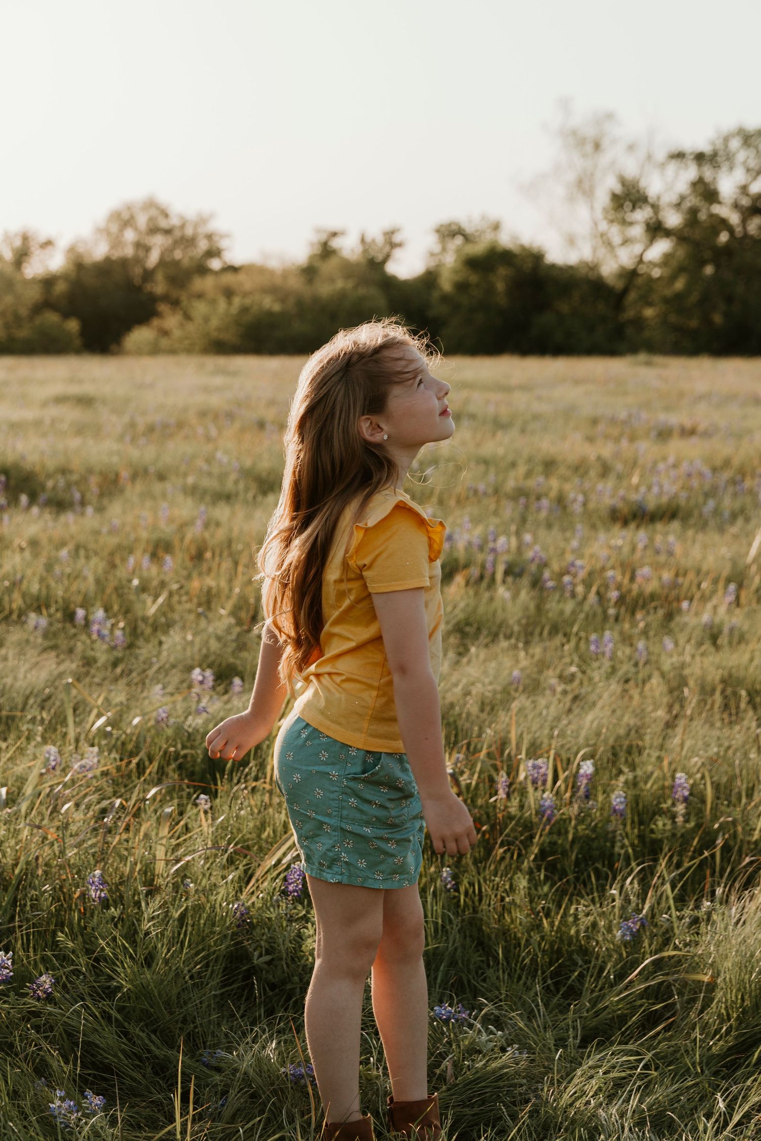Young person in yellow shirt and green shorts stands joyfully in a wildflower meadow at golden hour.