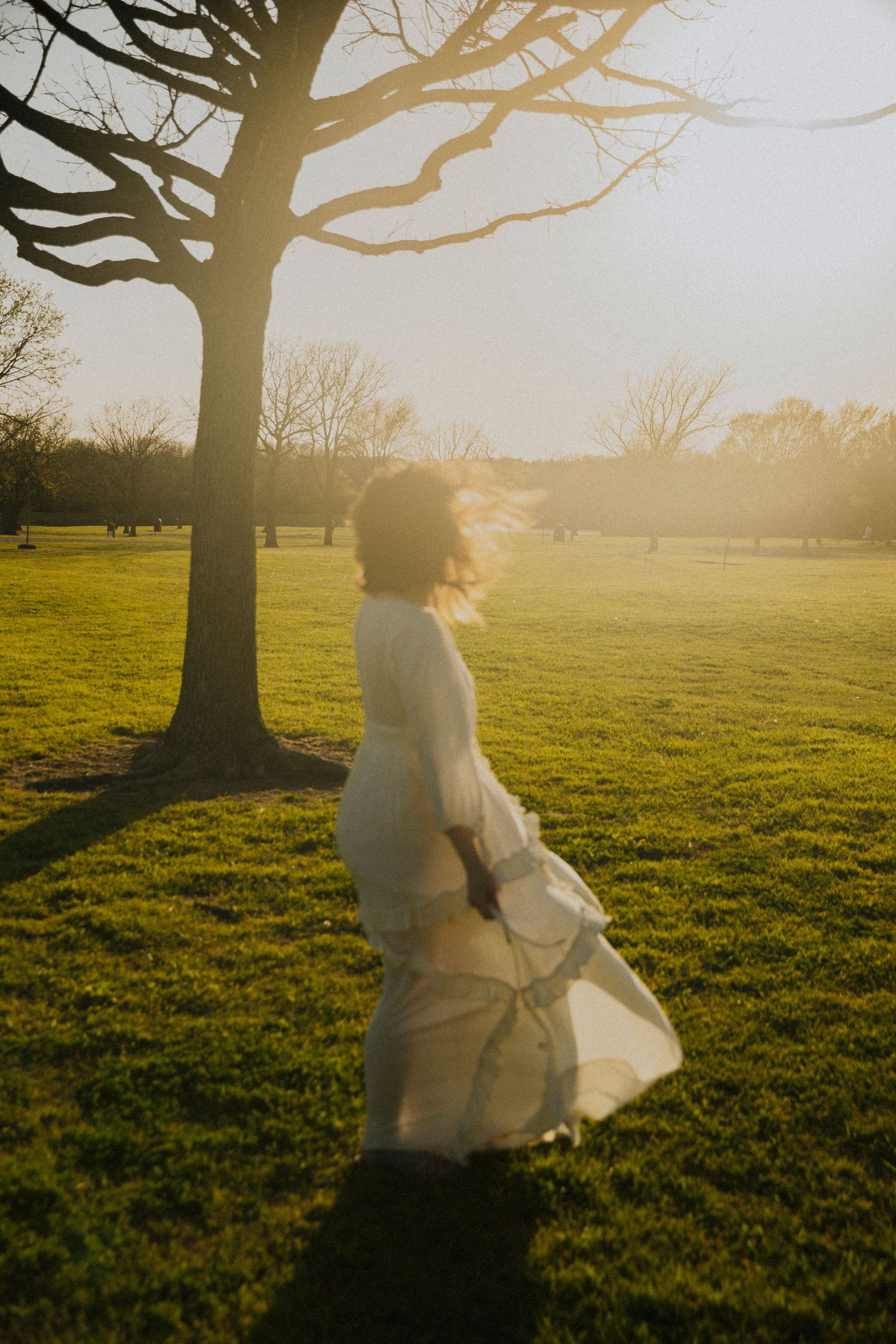 Ethereal figure in a flowing white dress stands by a tree during golden hour in a grassy park.