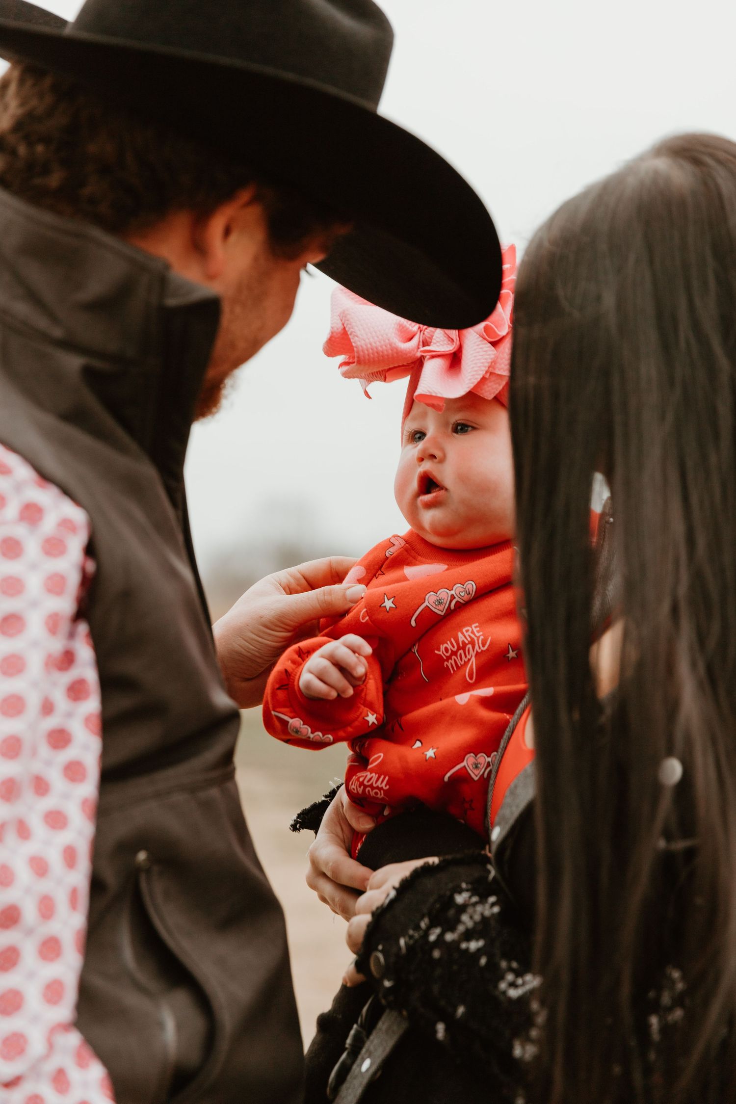 A baby in red clothing and pink bow being held during a family photoshoot in a rustic outdoor setting.