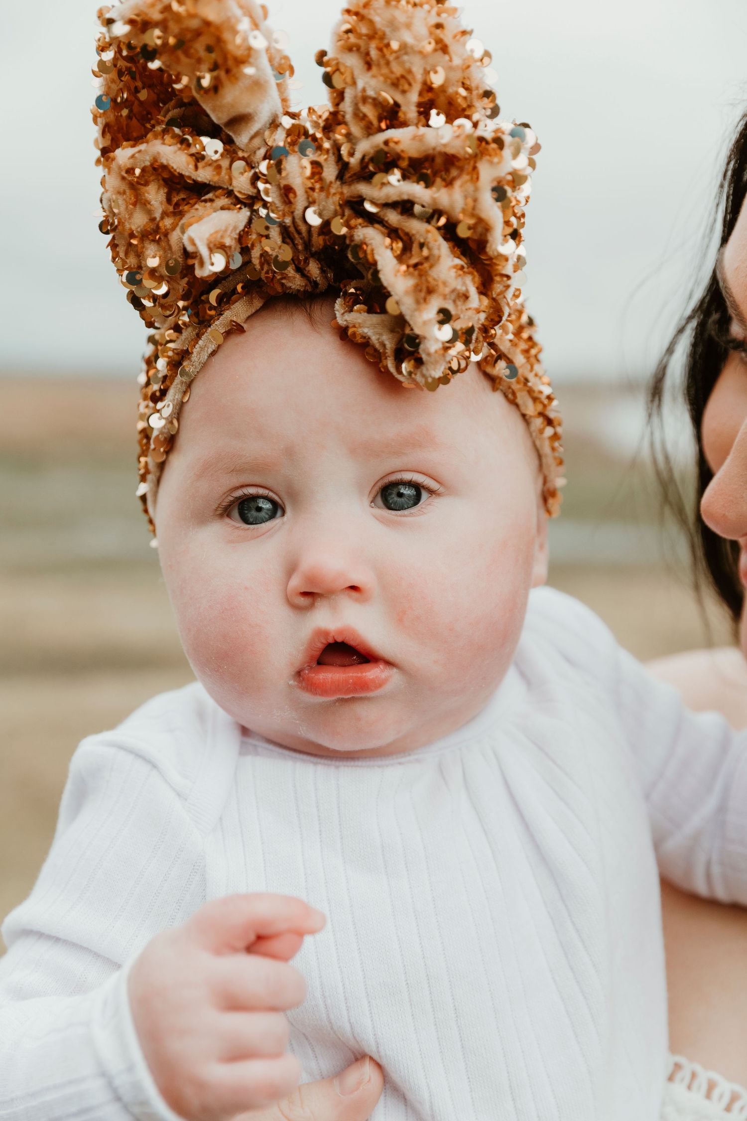 Baby wearing sparkly gold bow headband and white sweater looking at camera.