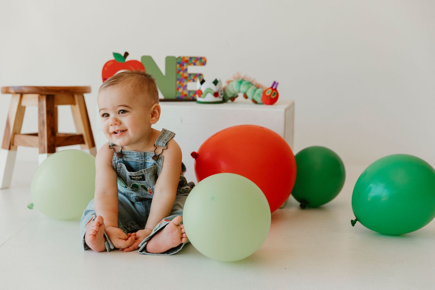 Baby in overalls smiling while sitting on floor with green and red balloons for a cake smash photo session.