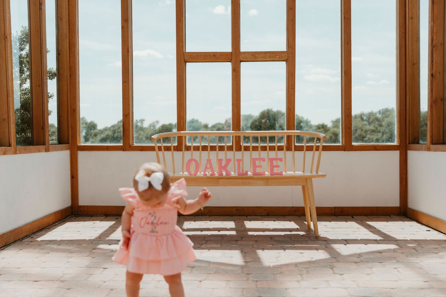Toddler girl in pink dress stands before a wooden bench spelling OAKLEE in a sunlit greenhouse with large windows.