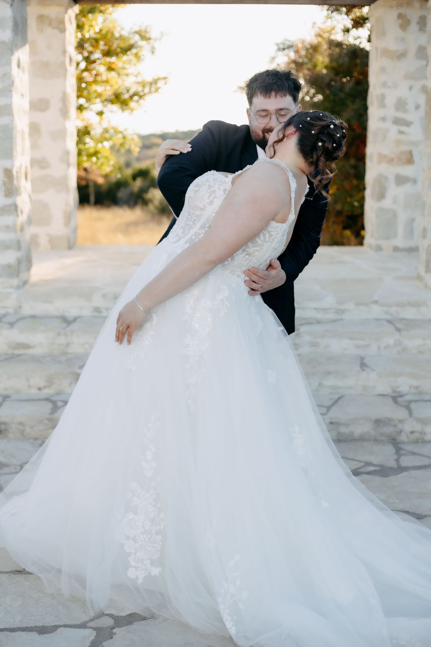A romantic wedding couple sharing a dip-style dance pose in front of stone pillars and columns at sunset.