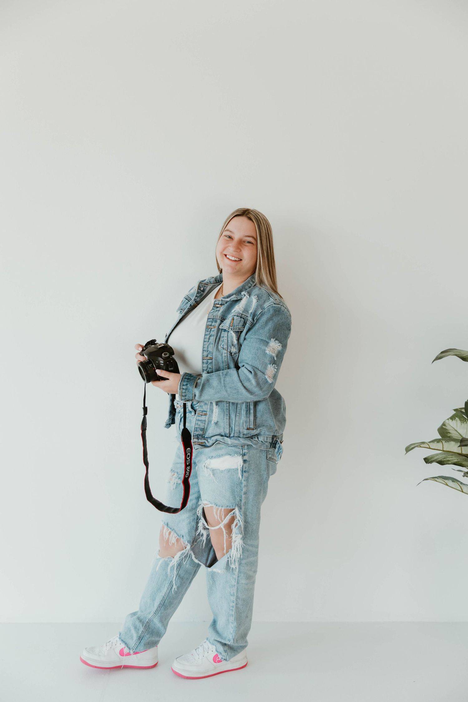 Person in denim outfit holding camera in bright studio setting with plant.