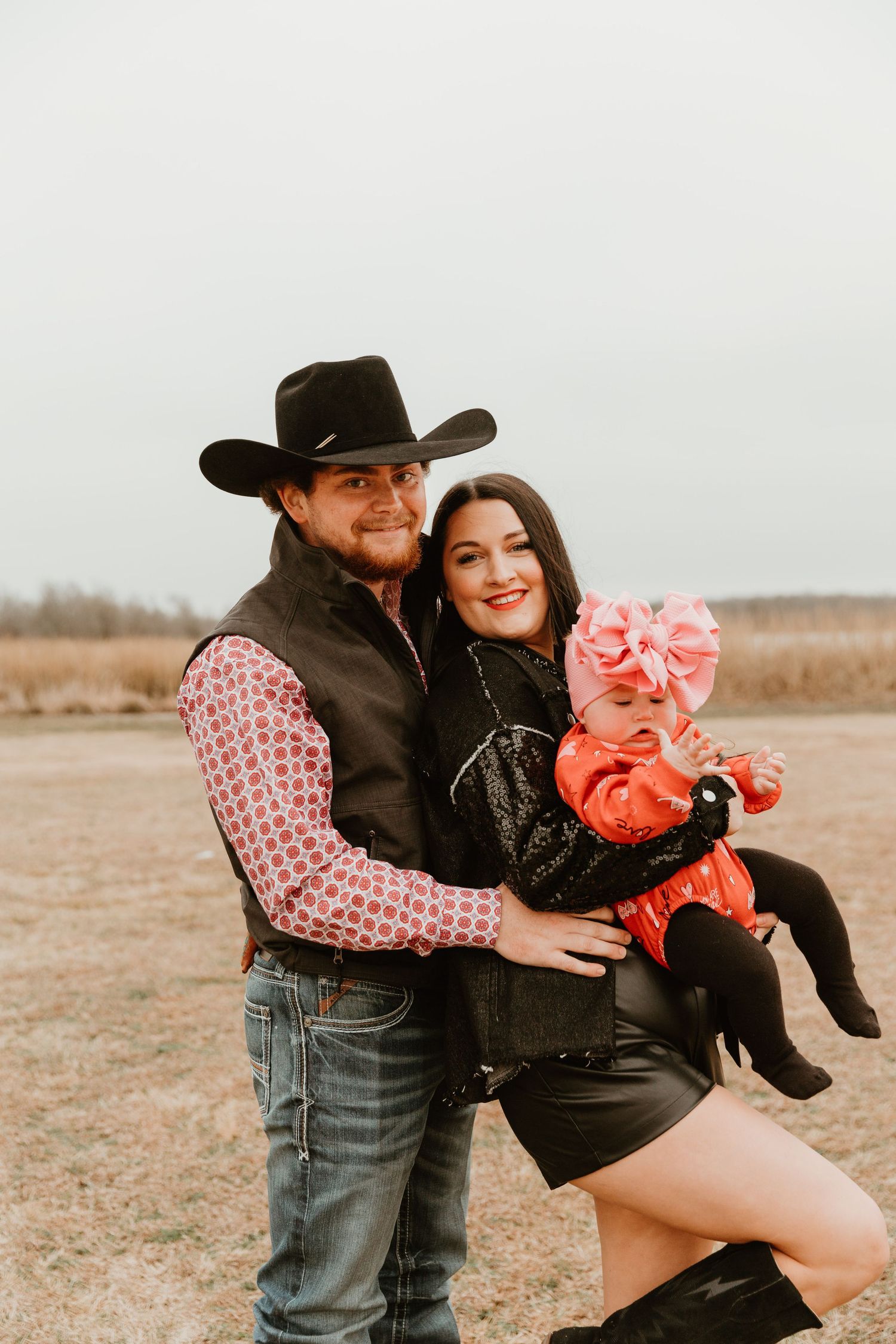 Family portrait in western attire with baby wearing red outfit and pink bow on open field.