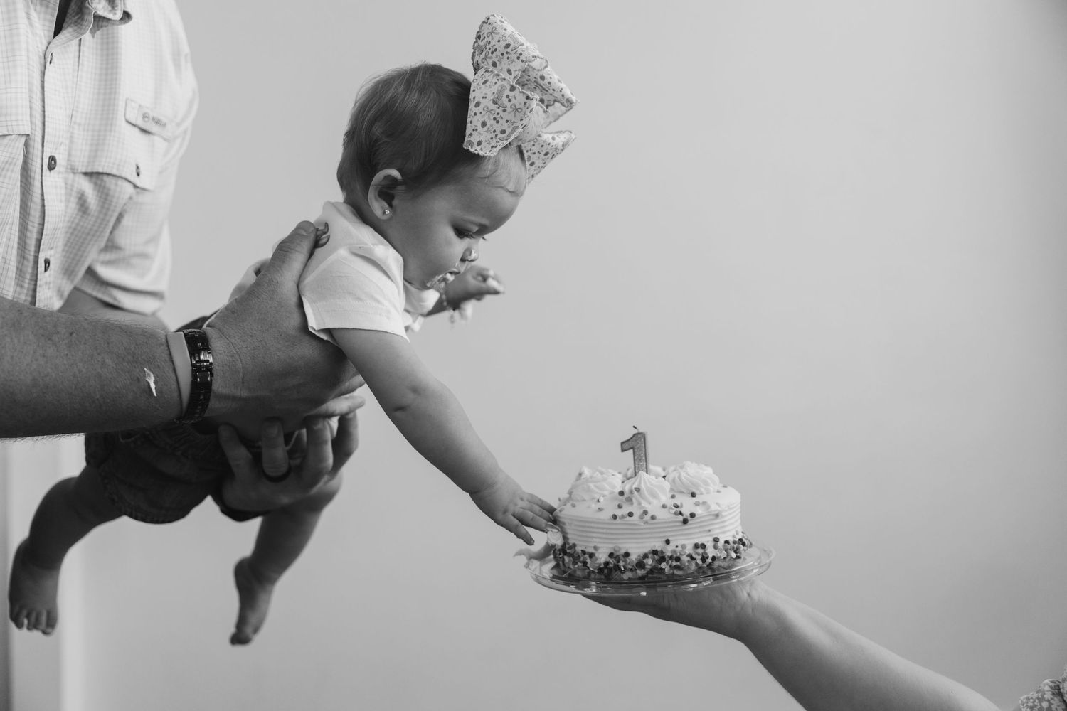 Black and white photo of a baby in a crown reaching for a first birthday smash cake held by an adult.
