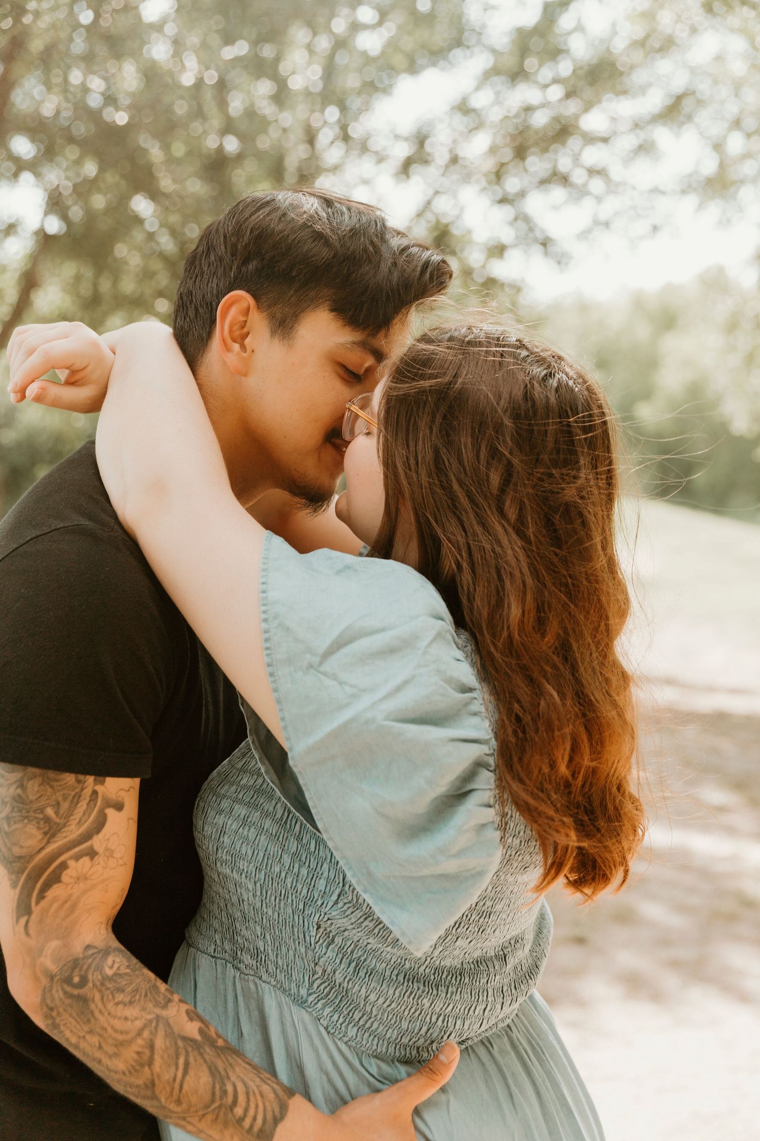 Romantic couple embrace outdoors with soft bokeh lighting and visible arm tattoo.