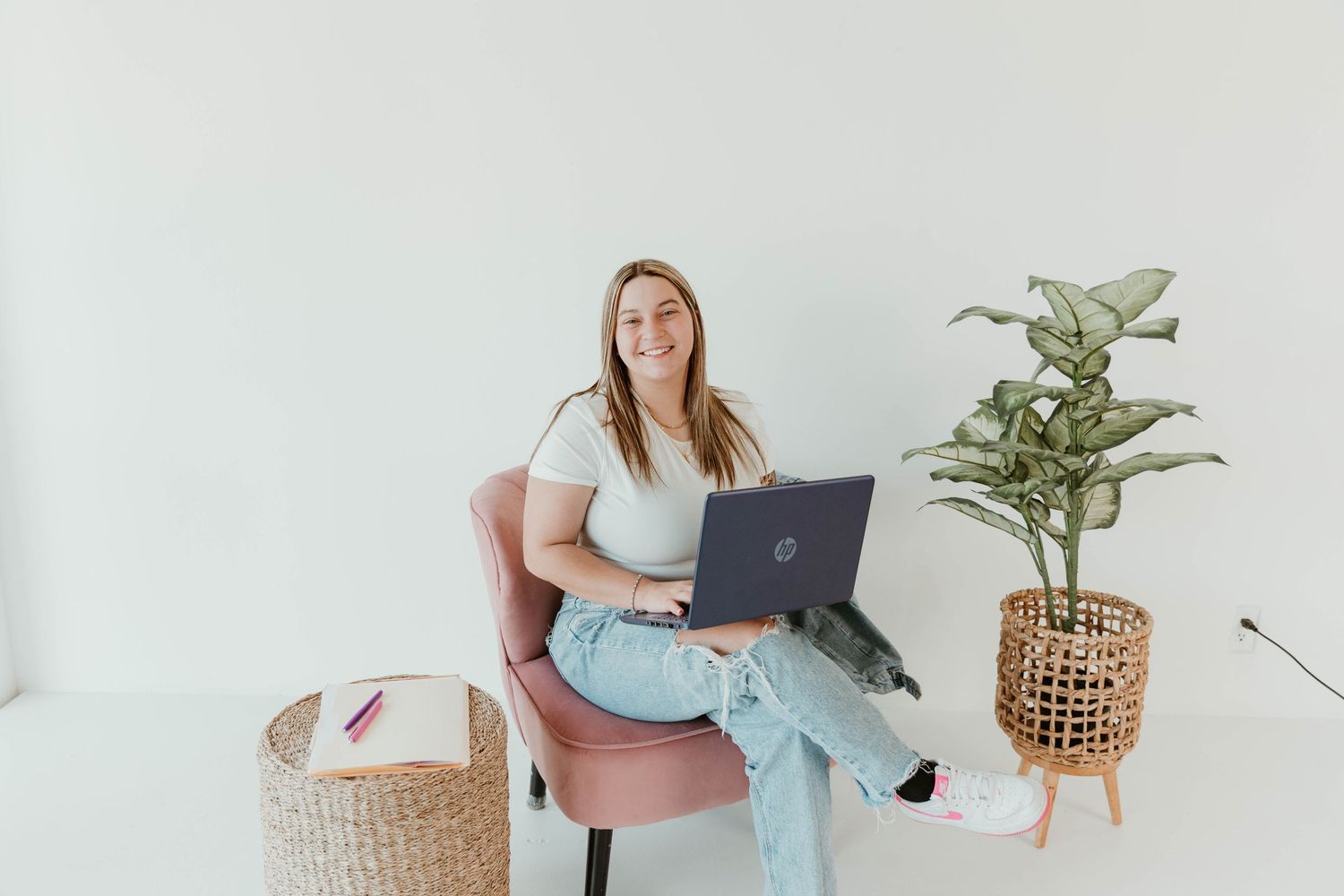 Person in casual wear working on laptop while sitting in pink chair with indoor plant nearby.