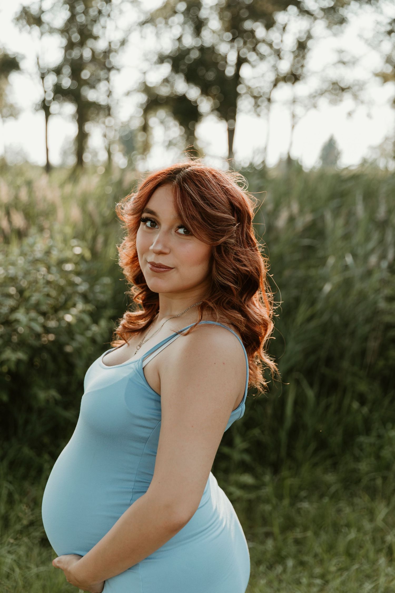 A summer maternity portrait showcases a flowing light blue dress against a natural outdoor backdrop.