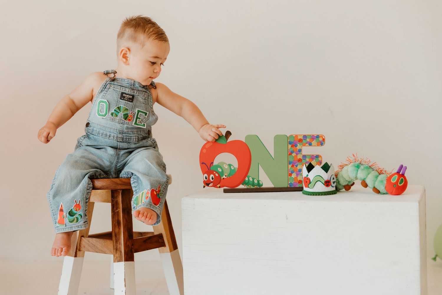 A baby in denim overalls sits on a wooden stool next to colorful nursery decor and toys.