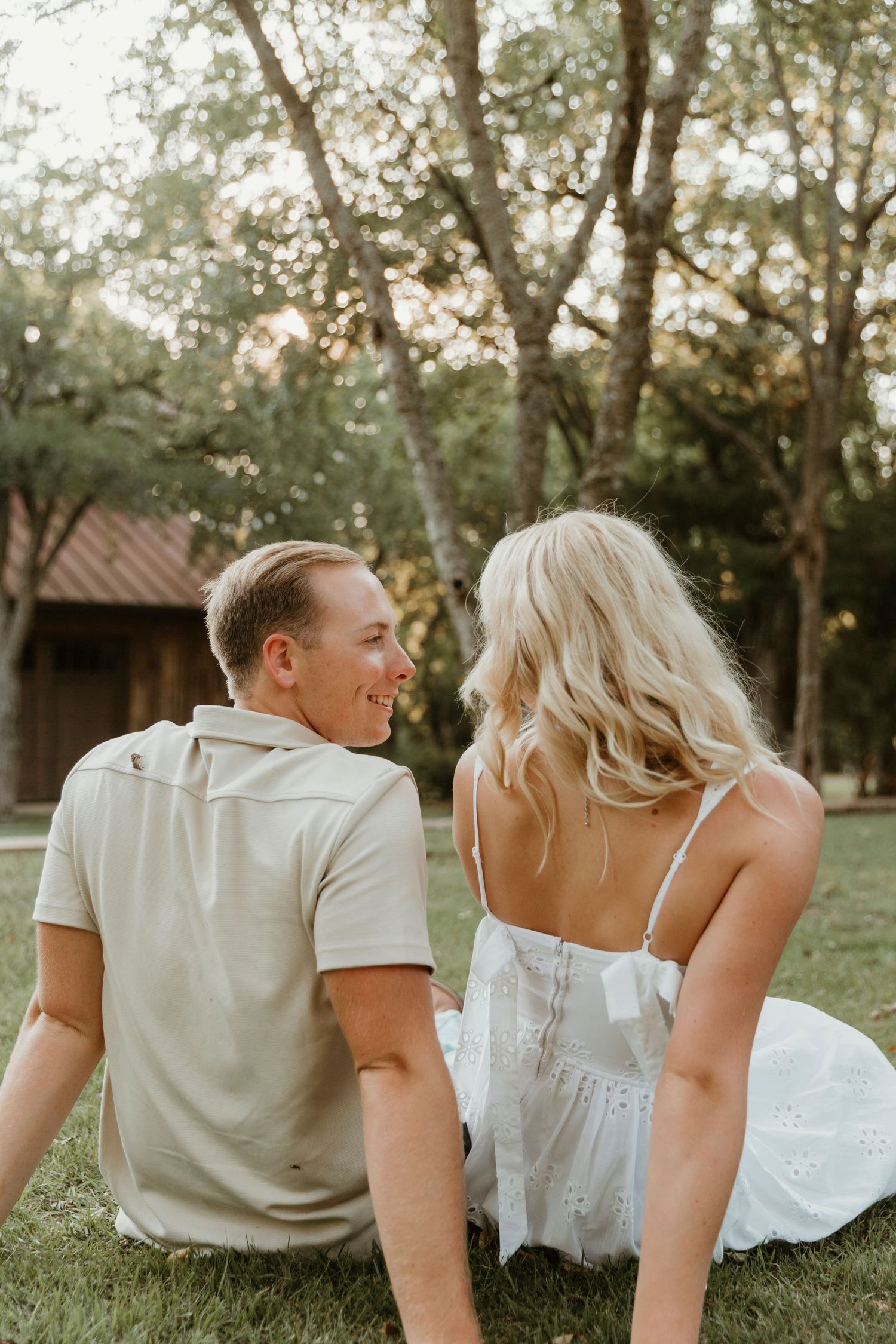 Romantic couple sitting on grass at sunset with rustic barn in background while sharing joyful moment together.