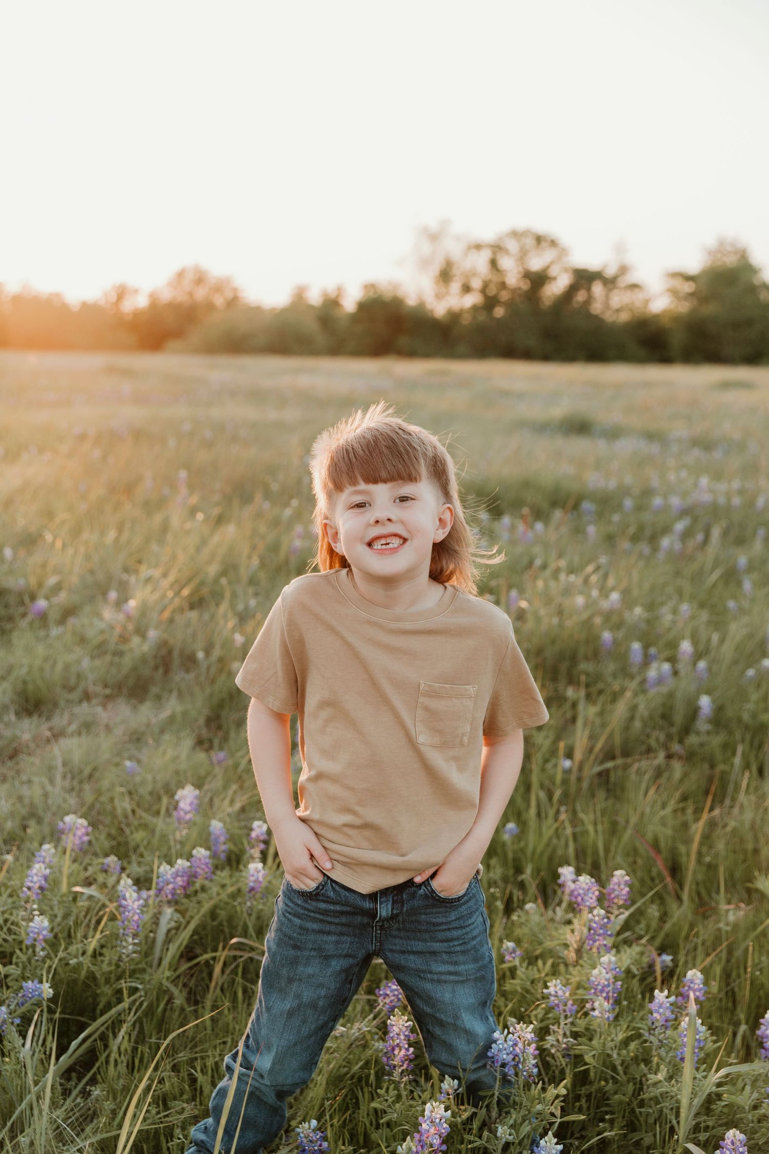Child in tan shirt posing playfully in wildflower field at sunset.
