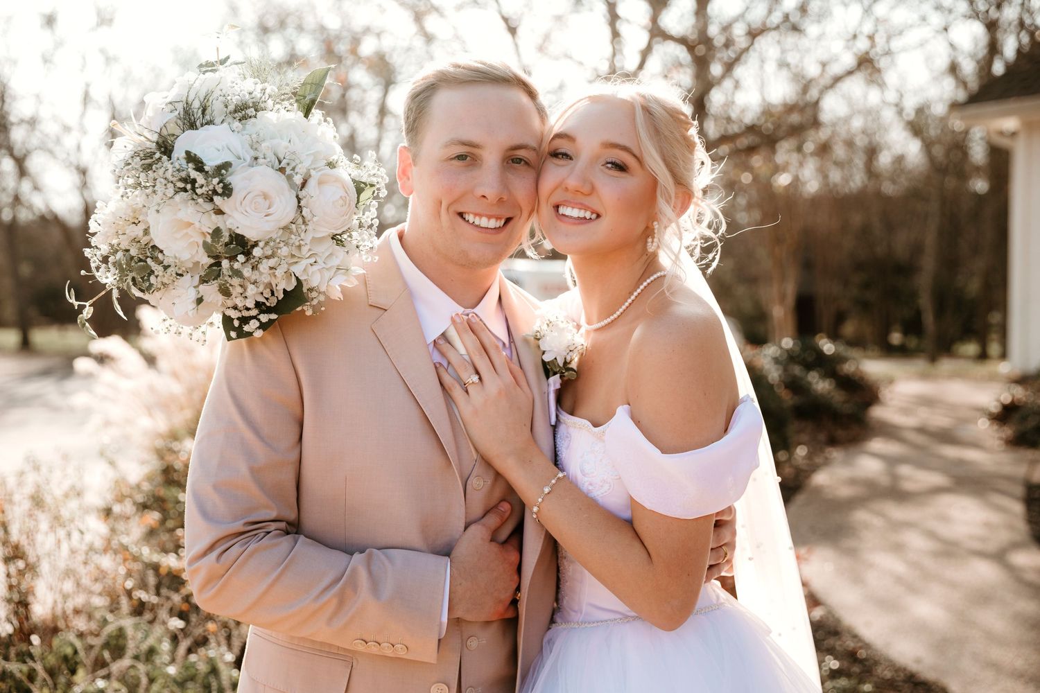 A romantic outdoor photoshoot featuring a couple in blush and white outfits embracing and smiling together in warm sunlight.