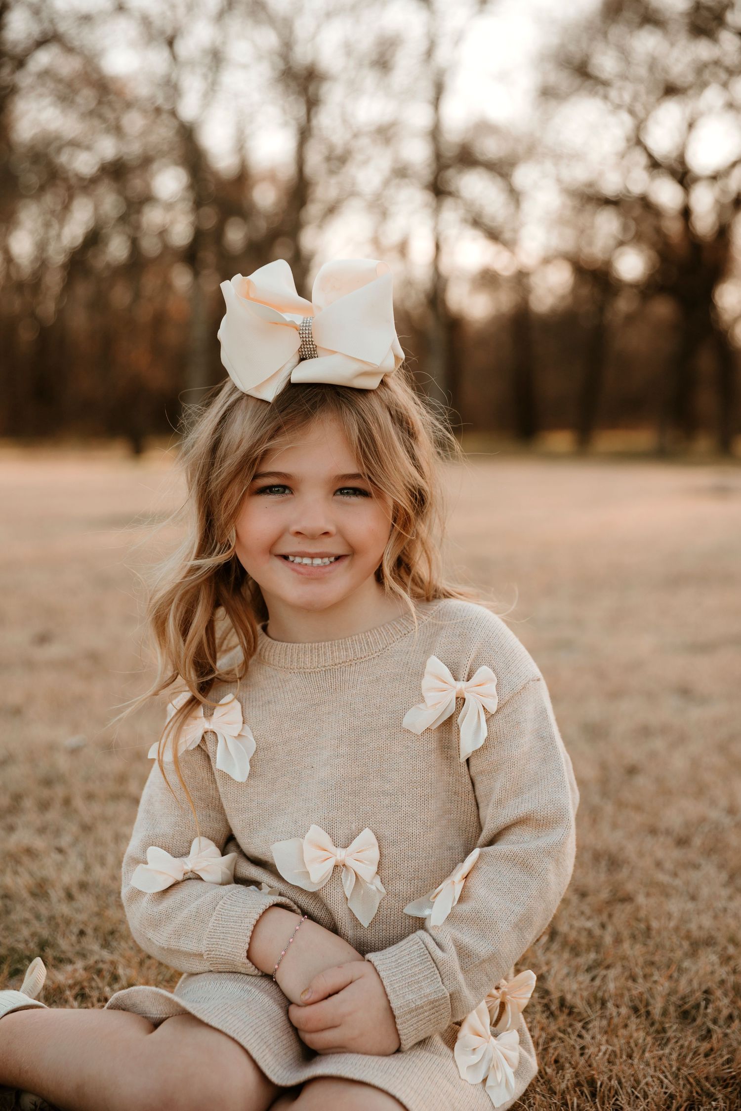 Child wearing cream sweater with bow details sits in autumn field during golden hour sunset.
