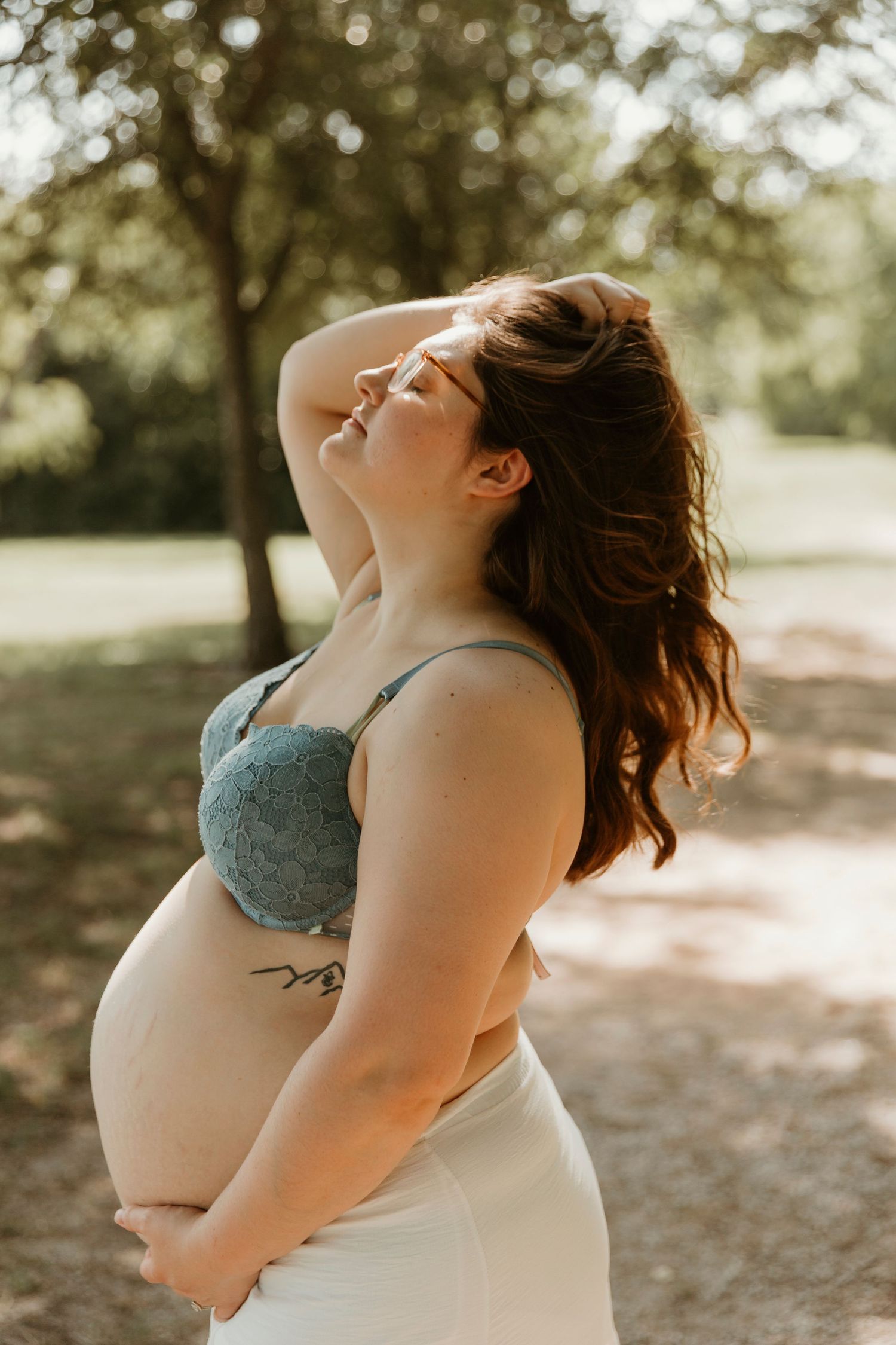 Side view of pregnant woman in teal dress posing outdoors with sunlit trees in background.
