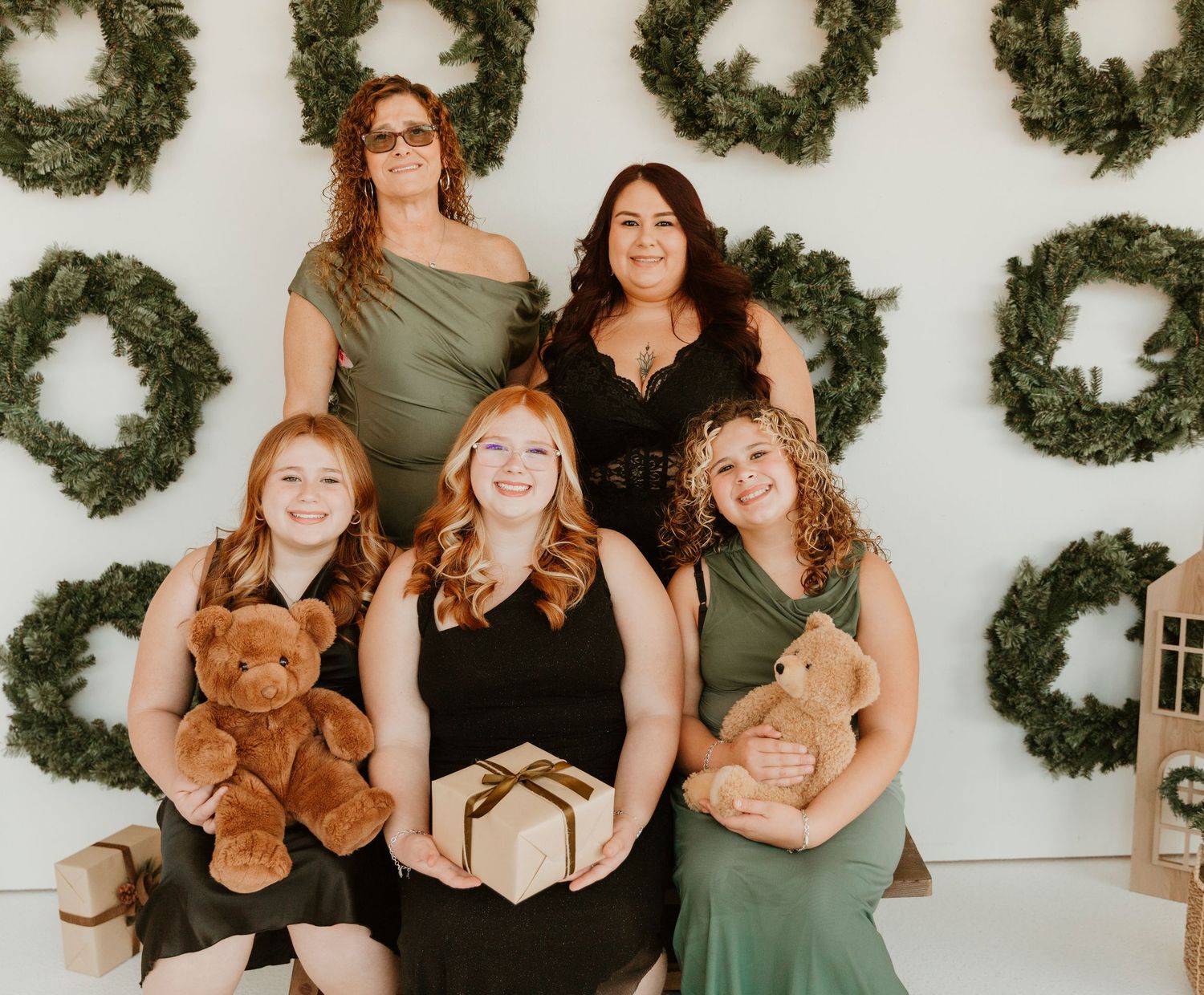 A group wearing black and sage green dresses poses against a white wall decorated with evergreen wreaths.