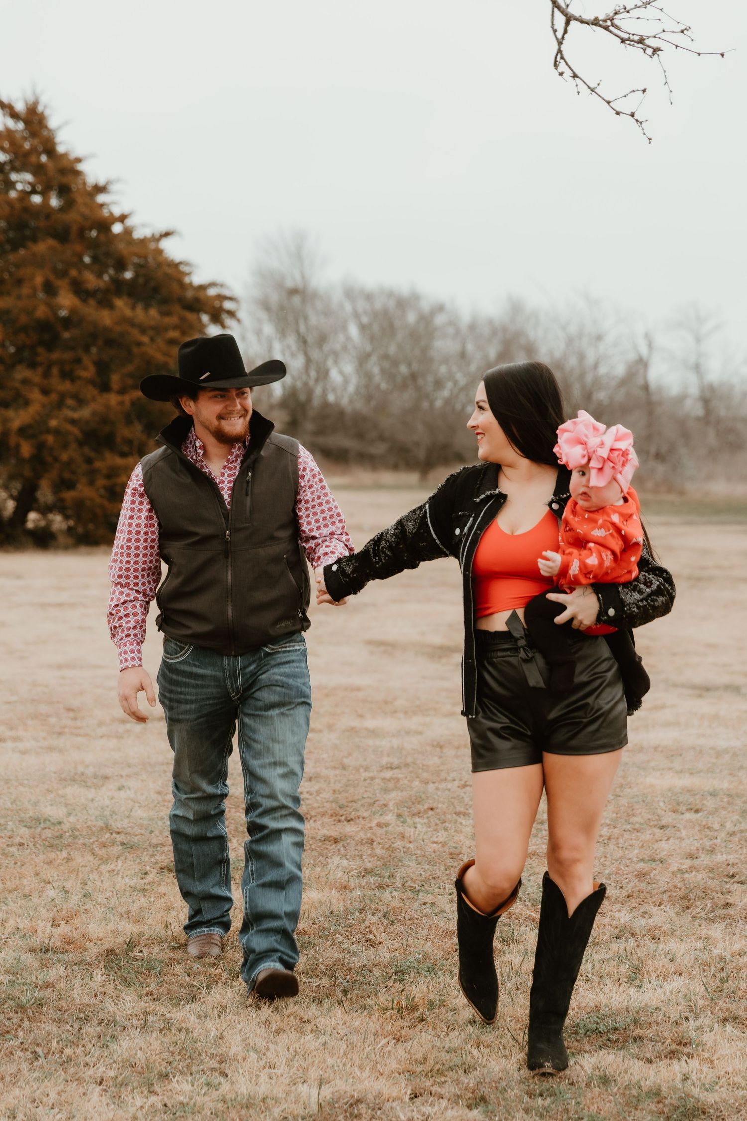 Couple in western wear walking together while holding baby in red outfit on winter field.