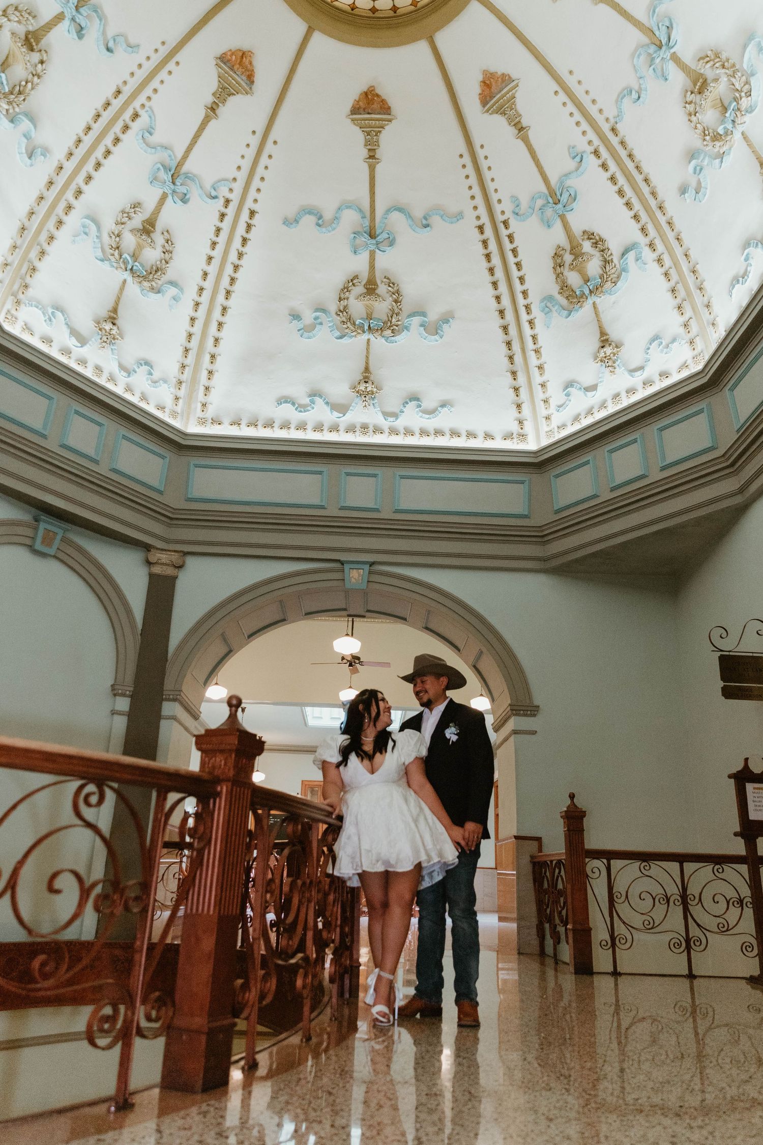 Wedding couple stands beneath ornate domed ceiling with decorative painting and archways.