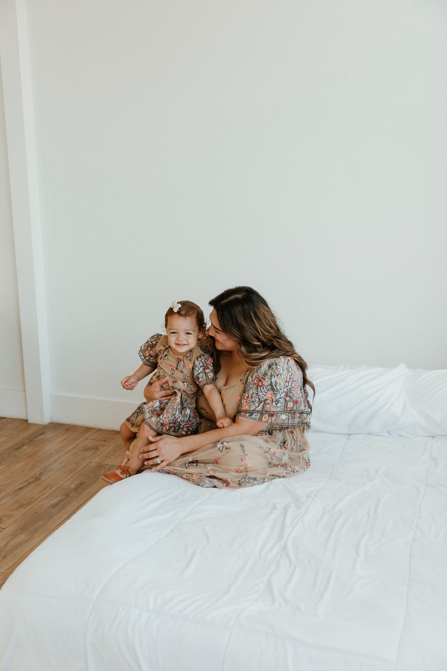 Mother and toddler sharing tender moment on white bed wearing matching floral dresses.