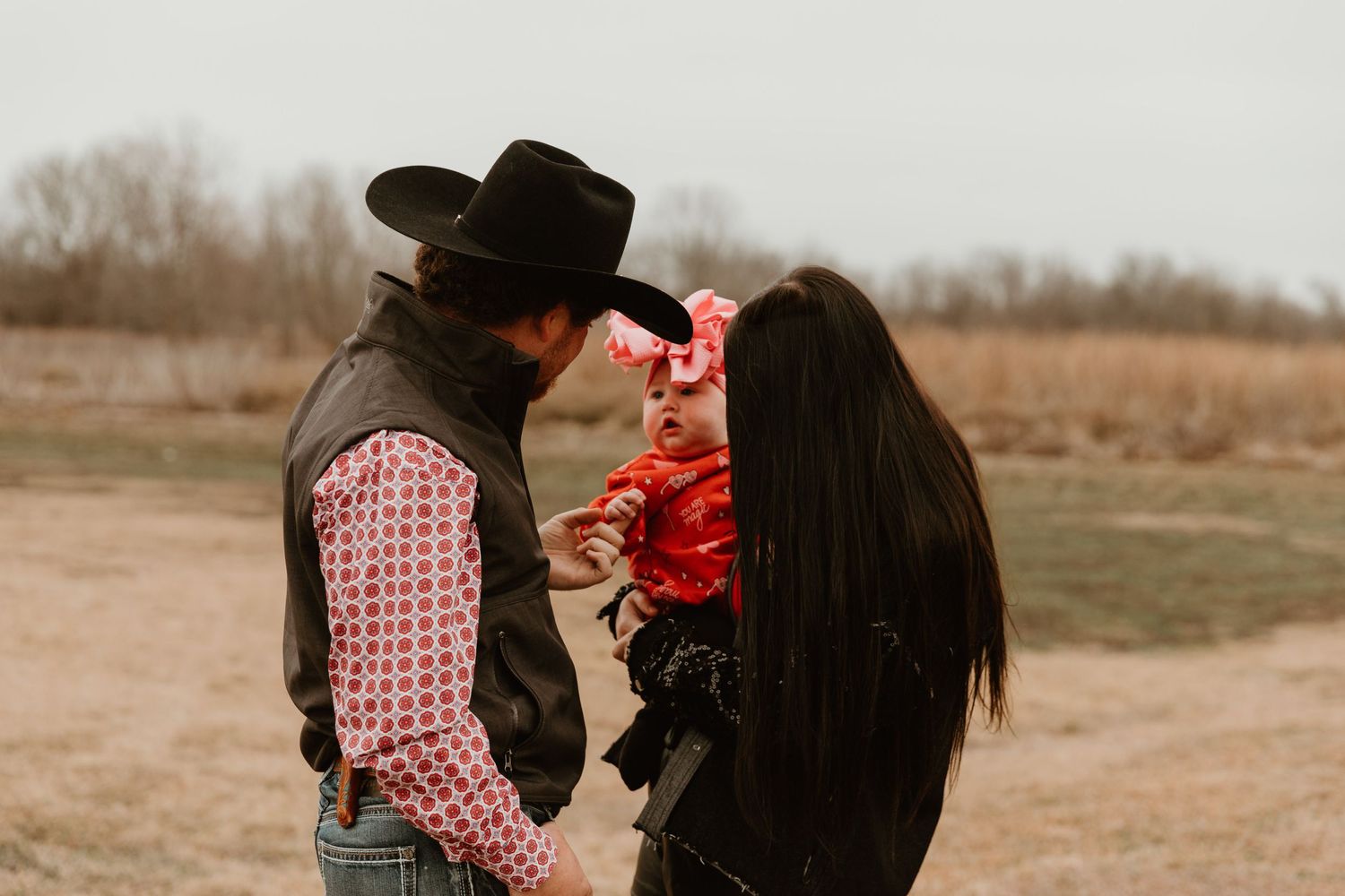 Intimate outdoor family portrait with baby in red outfit against winter landscape.