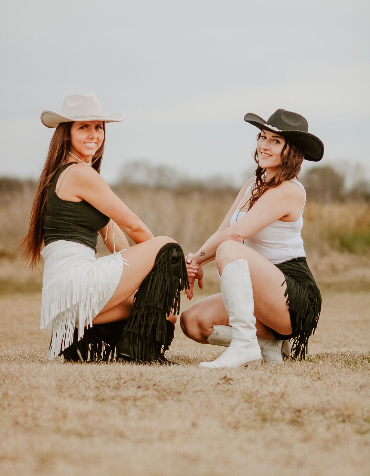 Two women in cowboy hats and fringe skirts sitting together in a field at sunset.