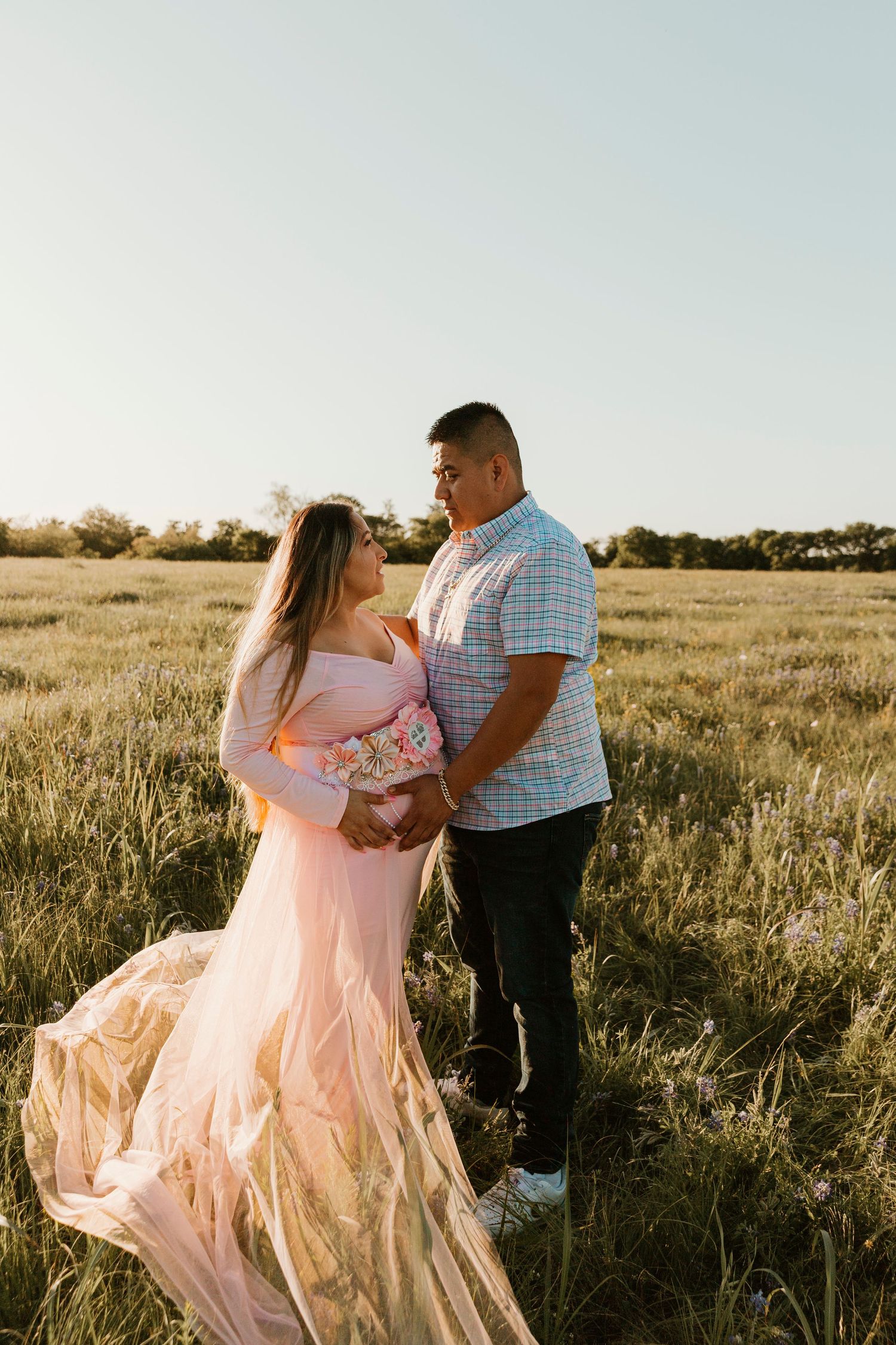Expectant couple in formal wear embracing in a sunlit field.